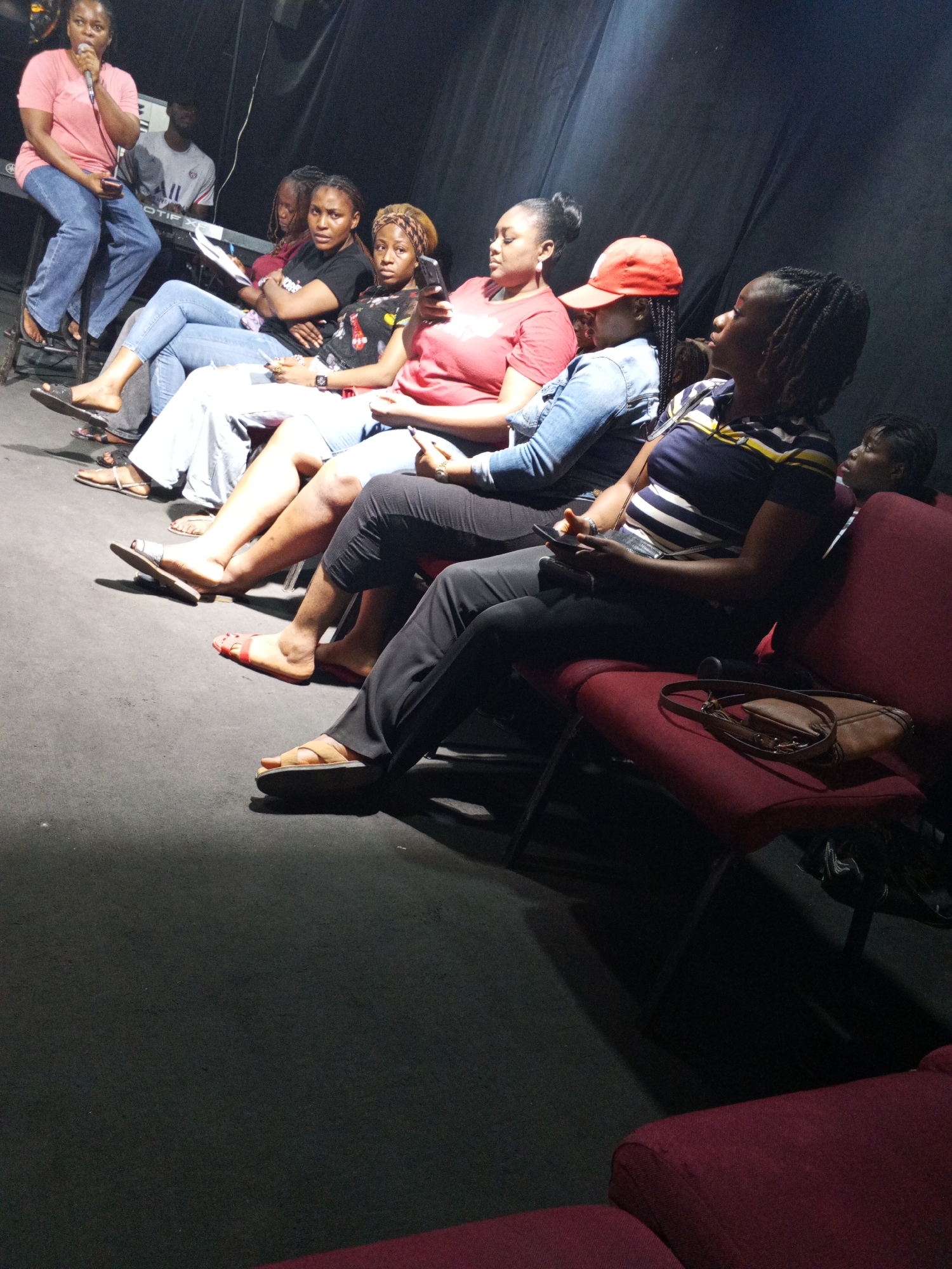 A group of people are seated in rows of chairs, likely in an indoor setting. A woman is seated on a stool at the front of the group, holding a microphone and speaking. Some individuals are wearing casual clothing, including jeans, t-shirts, and sandals. One person wears a red baseball cap. Some are looking towards the speaker. A purse sits on one of the chairs. In the background there appears to be a man seated at a keyboard. The location may be Abuja, Nigeria.