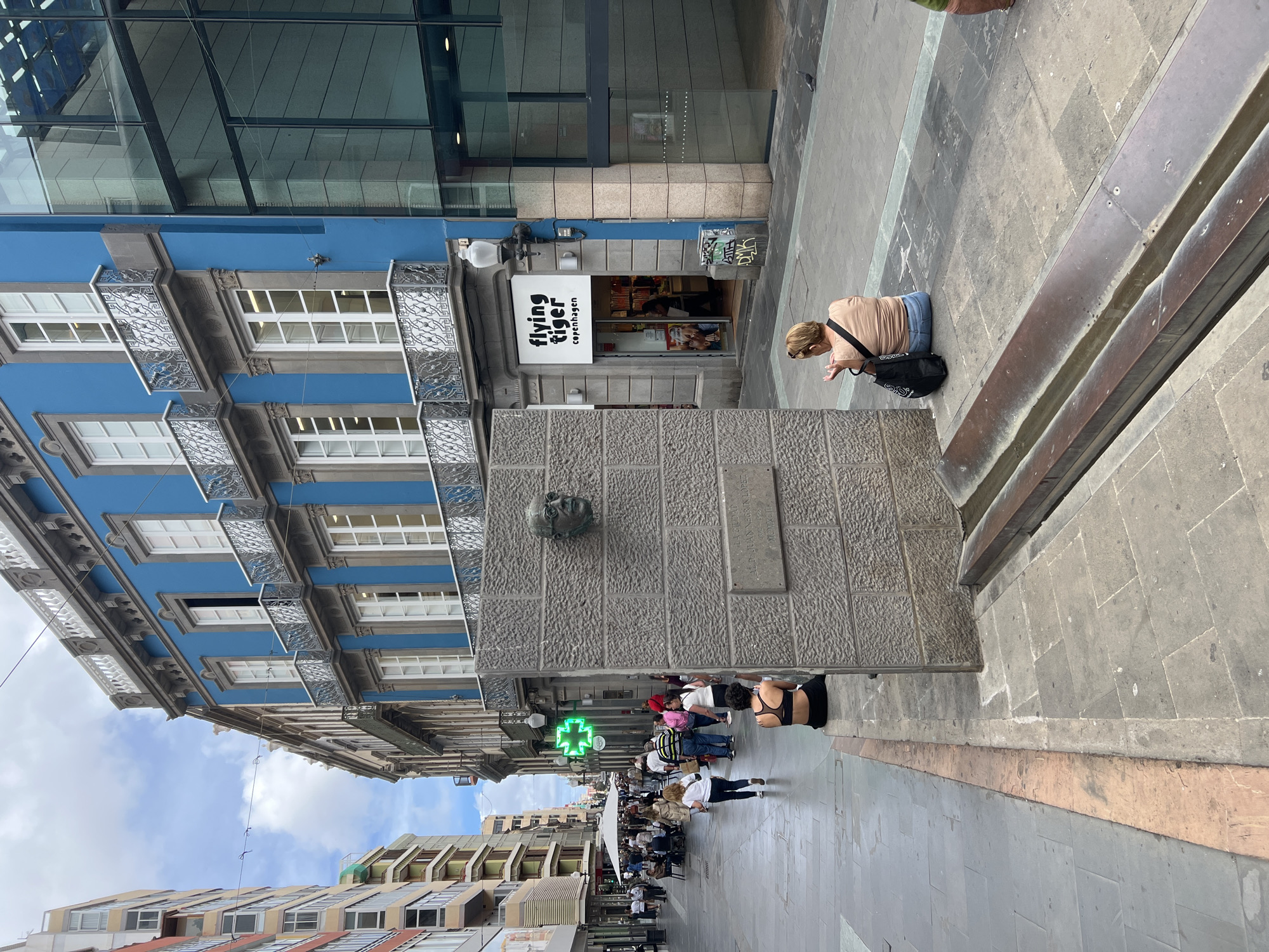 The image captures a street scene in Las Palmas de Gran Canaria, Spain. A four-story blue building with white-framed windows and decorative wrought iron balconies stands to the left of the frame, merging with a modern glass structure above. To the right of the building's ground level is a "Flying Tiger Copenhagen" store.
A large stone monument with a bronze head affixed to it is centrally positioned, acting as a focal point. People are visible on the sidewalk, including a woman sitting on the edge of a raised platform near the monument, appearing to use her phone. Pedestrians are walking further down the street, and a green pharmacy sign is visible in the distance. The sky is partly cloudy, and other buildings line the street.