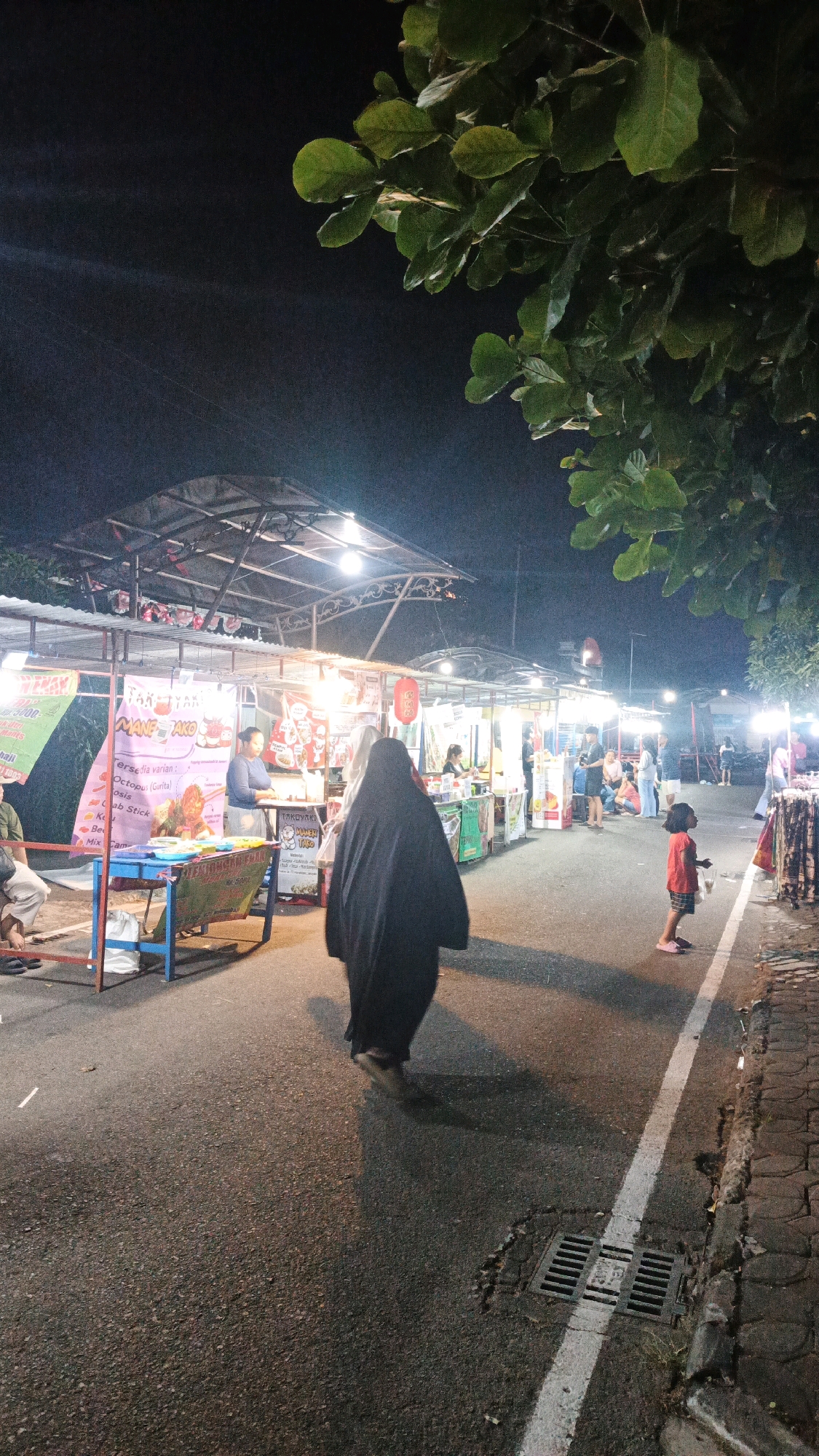 The image depicts a nighttime scene at a street food market in Surakarta, Indonesia. The market is lined with stalls under canopies, illuminated by artificial lights. Several people are visible, including a woman in a black head covering walking away from the camera, a child in a red shirt and pink shoes standing in the street, and vendors attending to their stalls. The market occupies a paved road, with a sidewalk constructed of interlocking bricks visible on the right side. A storm drain is present in the road near the foreground. Lush green foliage from a tree overhangs the top of the frame. Banners and signage on the stalls indicate food items and promotions. The overall atmosphere suggests a bustling evening market environment.