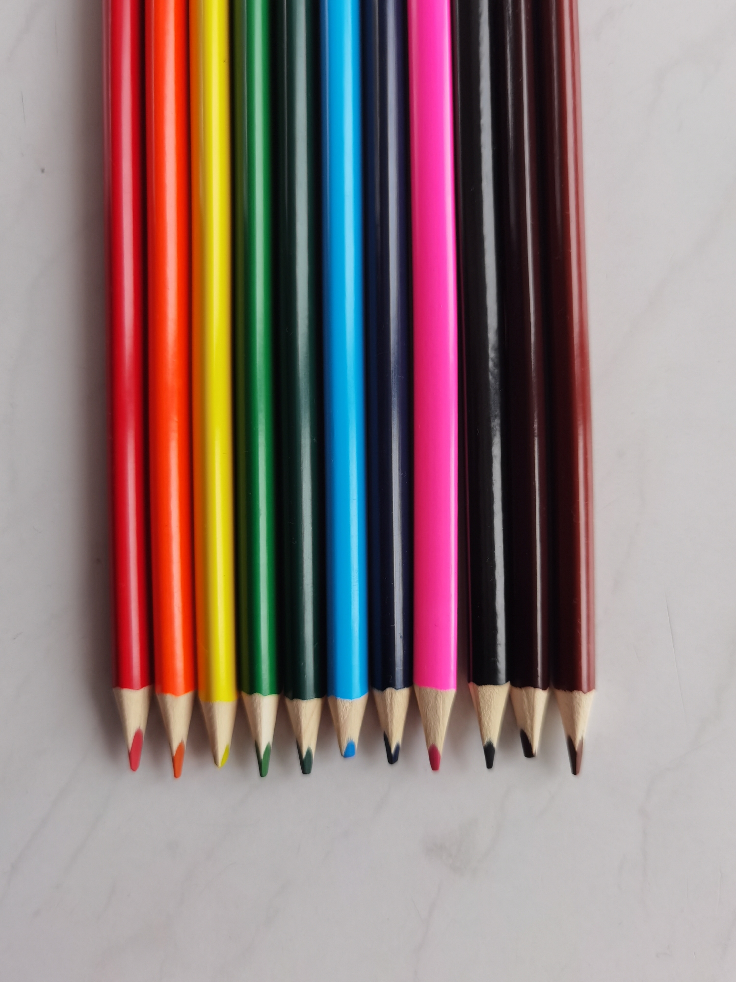 The image shows a row of twelve colored pencils on a white marble surface. The pencils are arranged in a slightly skewed line, with the tips facing down. The colors, from left to right, are red, orange, yellow, green, dark green, blue, navy blue, purple, pink, black, dark brown, and brown. The pencils are sharpened, and the tips are visible. The background is a white marble surface with subtle gray veins. No people, actions, or interactions are depicted. The environment is a plain surface. The image does not provide enough information to determine if this photo was taken in the city of Perm, Russia.