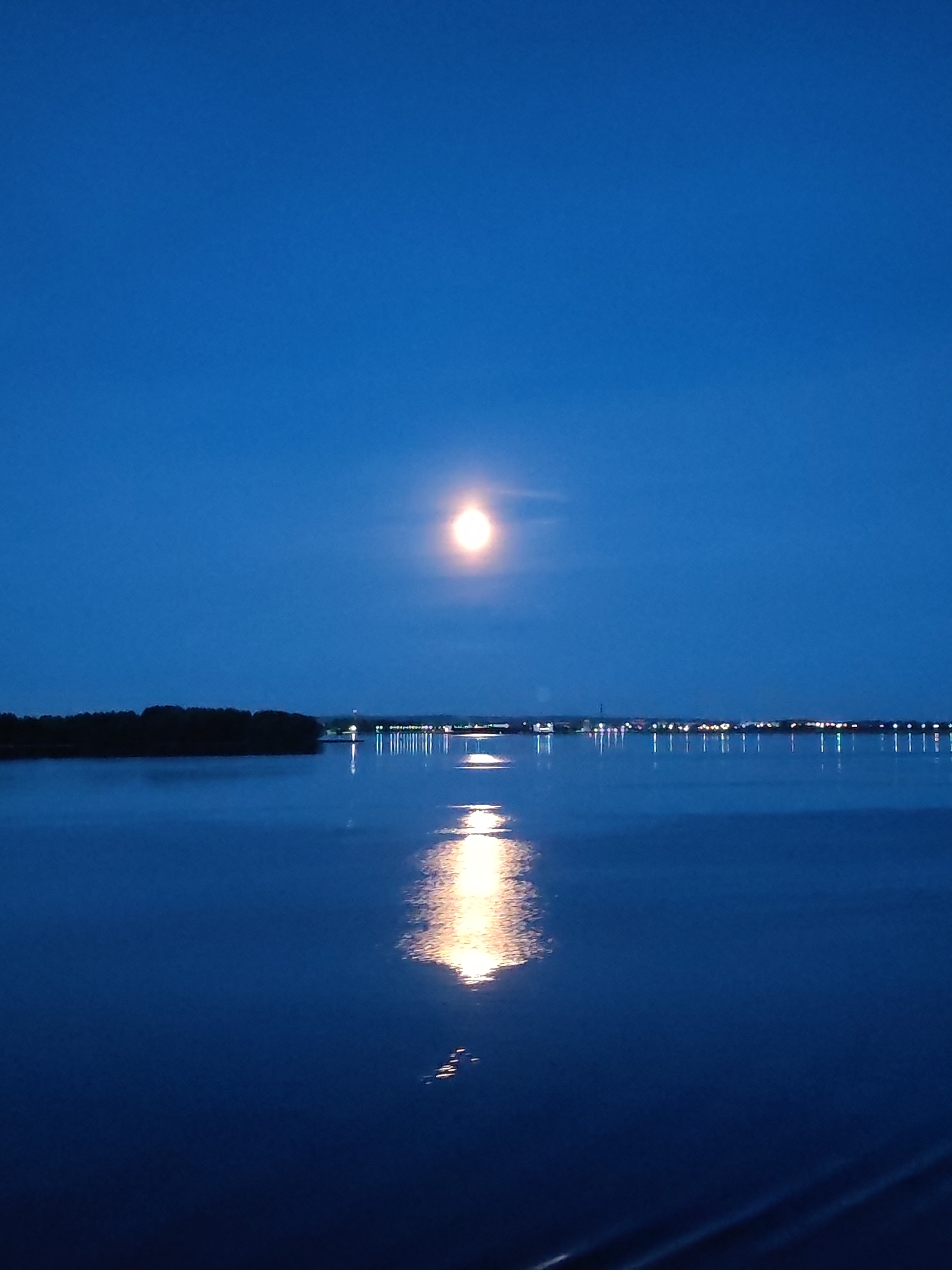 The image shows a body of water reflecting the moon and a distant shoreline with lights. The sky is a deep blue, indicating it is either dusk or night. On the left side of the image, there is a dark silhouette of land with trees. Across the water, the shoreline is lined with many small lights, suggesting a town or city. A bright moon is visible in the upper center of the sky, and its reflection creates a glowing path on the water's surface. The water itself is dark and calm. The image appears to have been taken from a vantage point looking across the water towards the moonlit landscape. The location is specified as Malinukha, Russia.