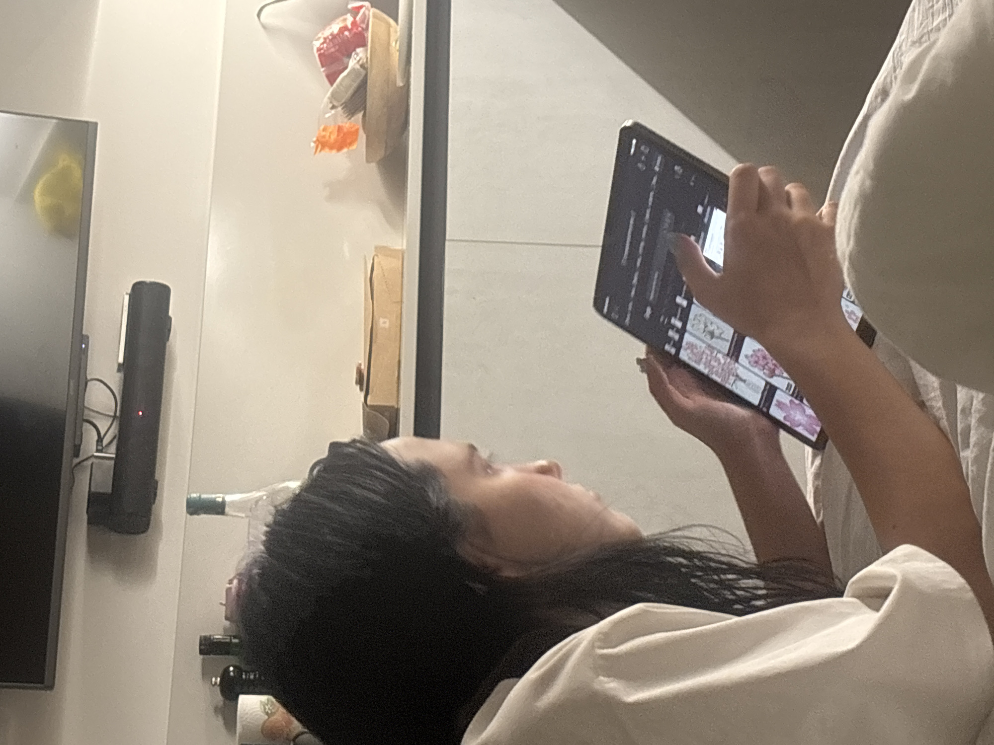 The image shows a person lying down and using a tablet computer. The person has long, dark hair and is wearing a white shirt. The tablet screen displays various images. In the background, there is a television mounted on the wall above a soundbar, and a shelf with items including a bottle and a bowl of what appears to be dried oranges. The scene is indoors, likely in a domestic setting. There is no discernible information in the image to suggest the location is in Cupra Marittima, Italy.