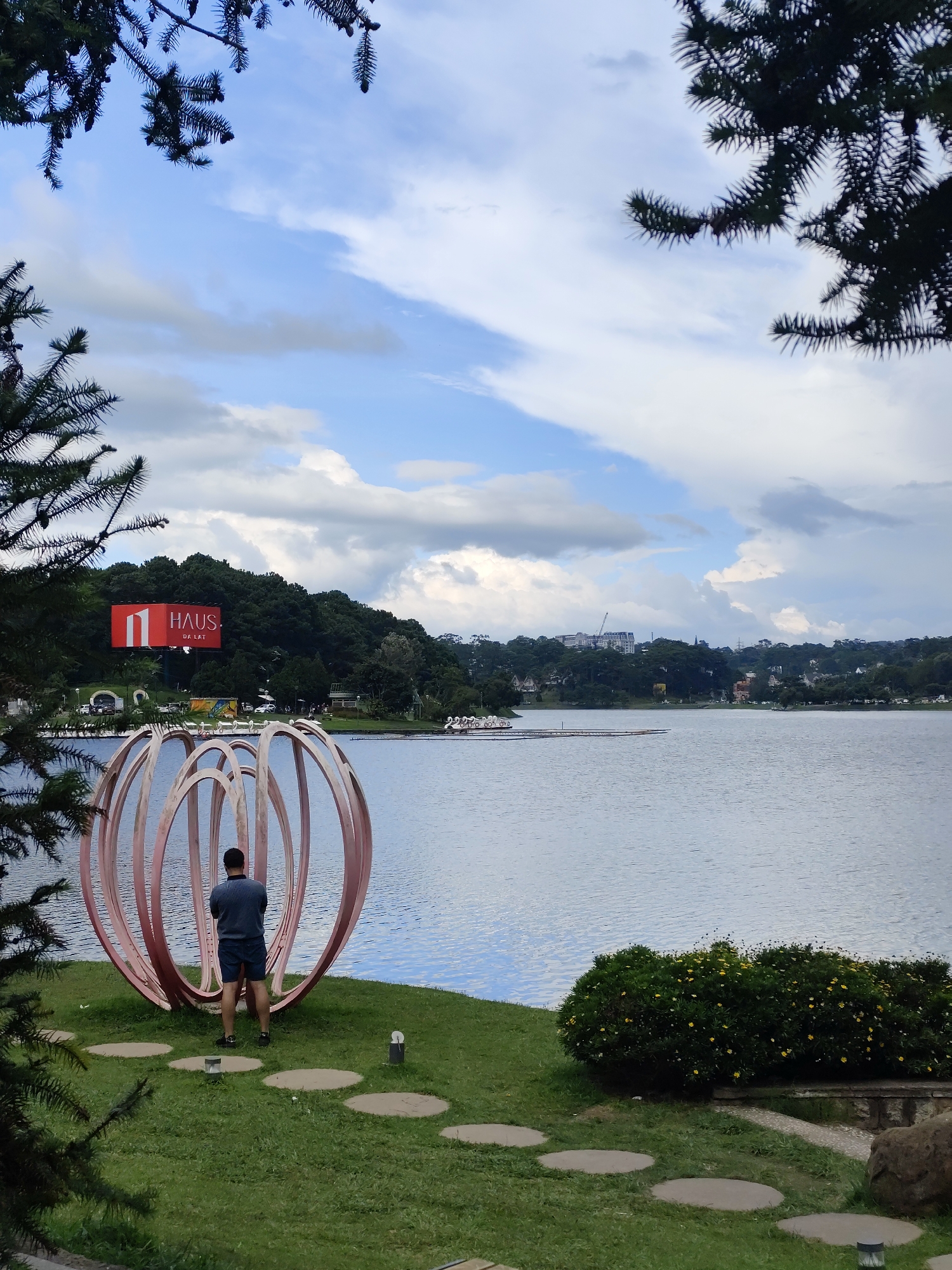 The image depicts a scenic view of Xuan Huong Lake in Da Lat, Vietnam. In the foreground, a man stands facing away from the camera, partially obscured by a large, spherical art installation with concentric pink rings. The installation is situated on a grassy knoll dotted with circular stepping stones and small garden lights. To the right, a dense green bush with yellow flowers adds a splash of color.
Across the tranquil blue water of the lake, the opposite shore is lined with a lush green treeline, behind which buildings and infrastructure are visible. A prominent red billboard with the text "HAUS" and "DK LAT" stands out against the foliage. Further in the distance, a construction crane indicates ongoing development. The sky above is a mix of blue and dramatic, cloudy formations, with the dark branches of pine trees framing the top of the image. A few paddle boats are visible on the lake in the middle ground, suggesting recreational activity. The overall impression is one of a peaceful, picturesque urban landscape.