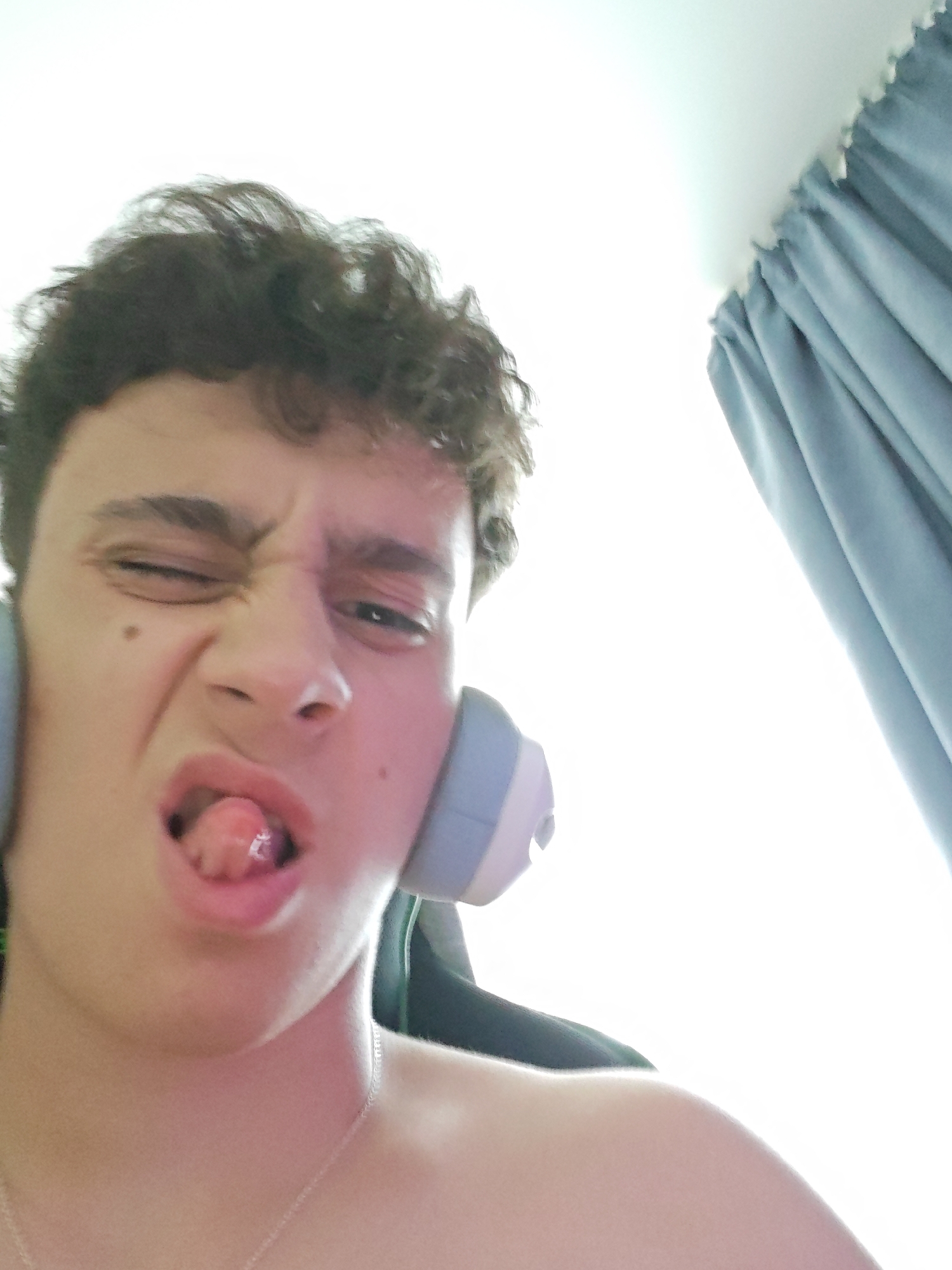 A young person with curly brown hair, wearing headphones and a necklace, sticks out their tongue and squints one eye at the camera. They are in what appears to be a brightly lit room, possibly with a window covered by blue curtains to the right. A gaming chair is visible behind them.