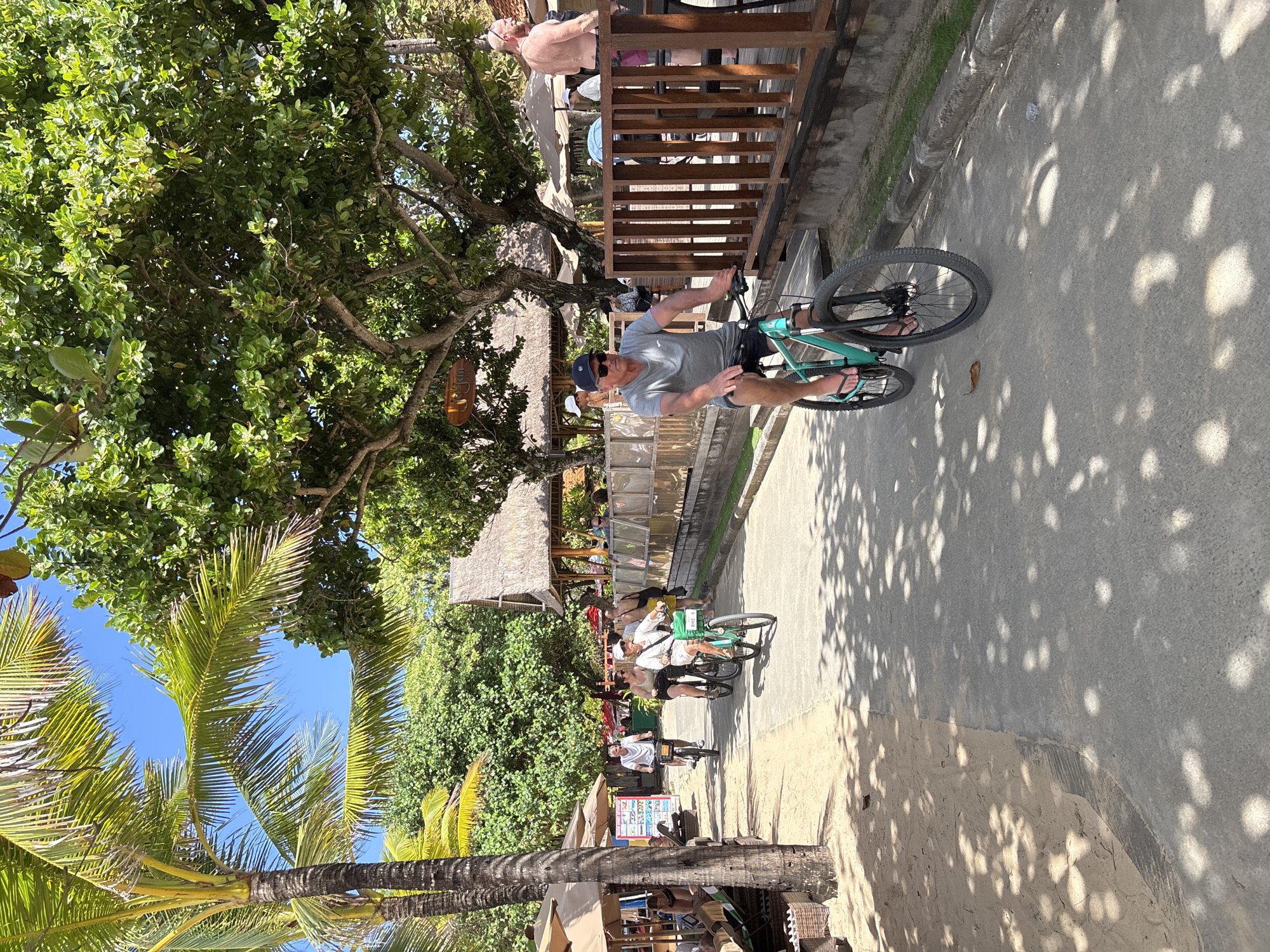 In Kuta Selatan, Indonesia, a group of people are cycling along a paved pathway beside a sandy beach. Palm trees and lush green foliage line the pathway, with thatched-roof structures and umbrellas visible in the background, suggesting a tropical, resort-like environment. The main subject in the foreground is a man wearing a grey t-shirt, black shorts, a baseball cap, and sunglasses, riding a teal-colored bicycle. He is facing towards the camera. Behind him, two women and another man are also on bicycles, moving in the same direction. One woman is wearing white, and the other is in a light-colored top and dark shorts. The man behind them is wearing a white t-shirt. Further down the path, more individuals and structures are visible, including a shirtless man with a light-colored hat standing near a wooden railing. The sunlight casts dappled shadows from the trees onto the pathway and the sand.
