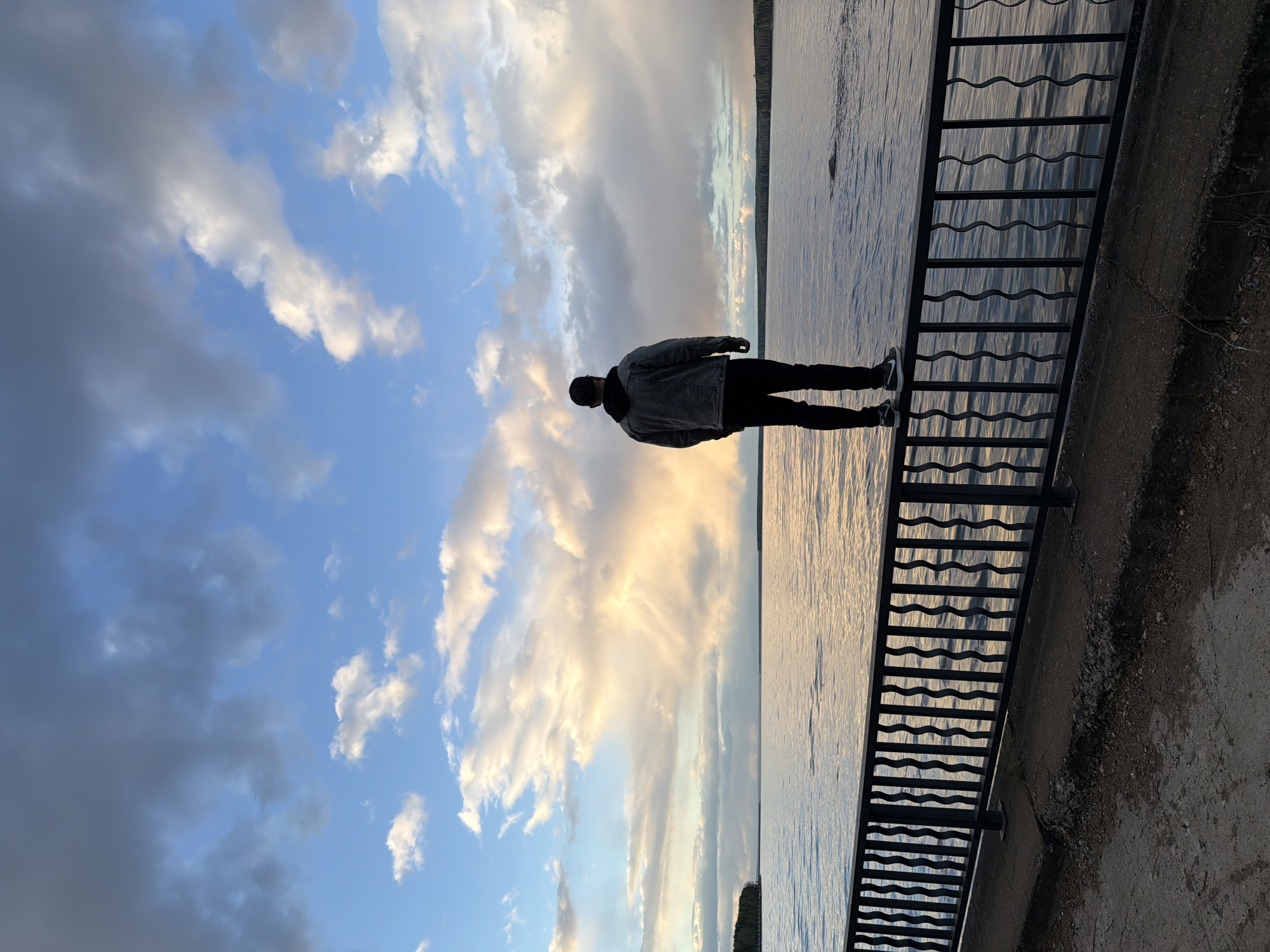 A man stands on a metal staircase next to a body of water, with a cloudy sky in the background. The location is Chaikovsky, Russia. The man is wearing dark pants and a jacket, and a hat. The sky is blue with white and gray clouds. The body of water appears to be calm and reflective. A concrete embankment is adjacent to the staircase. The image is oriented on its side.