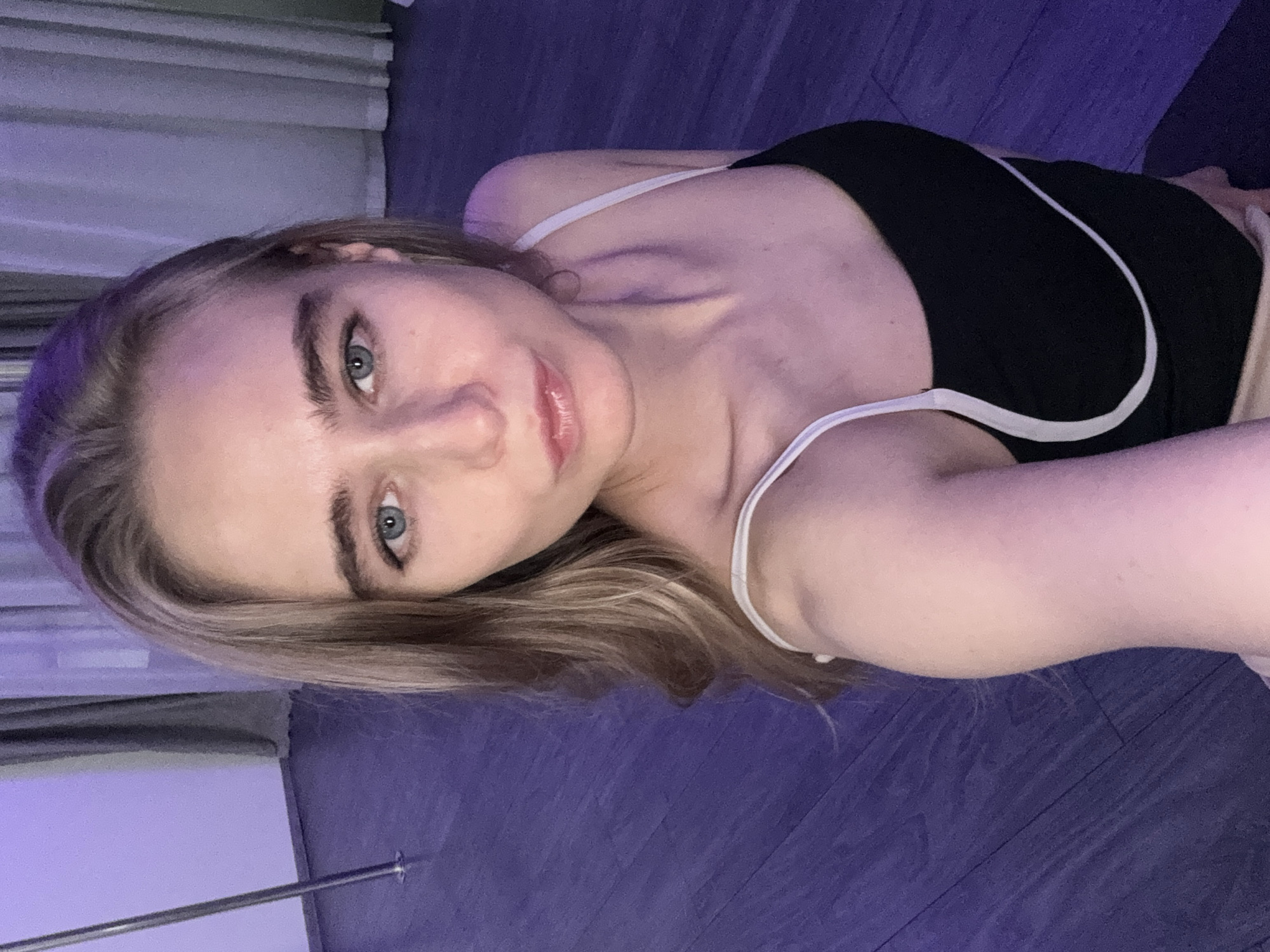 A young woman with light-colored, long hair and blue eyes is the central subject of the image. She is lying on her side, facing the viewer, with her head slightly tilted. She is wearing a black top or camisole with thin white straps and white piping. Her expression is calm, featuring a soft, gentle smile and slightly parted lips.
The setting appears to be indoors. She is positioned on a dark-colored floor that resembles wood, with visible grain. In the background on the left, there are curtains or blinds, and a slender, silver-colored metal pole stands vertically, extending from the bottom to near the top of the frame. The scene is illuminated by strong purple or violet ambient lighting, which casts a distinct hue over the floor, walls, and the subject's hair and skin. This artificial lighting creates a moody and atmospheric ambiance.
The activity taking place is a relaxed pose for a photograph, likely a selfie given the angle and proximity. There is no visible text in the image.
