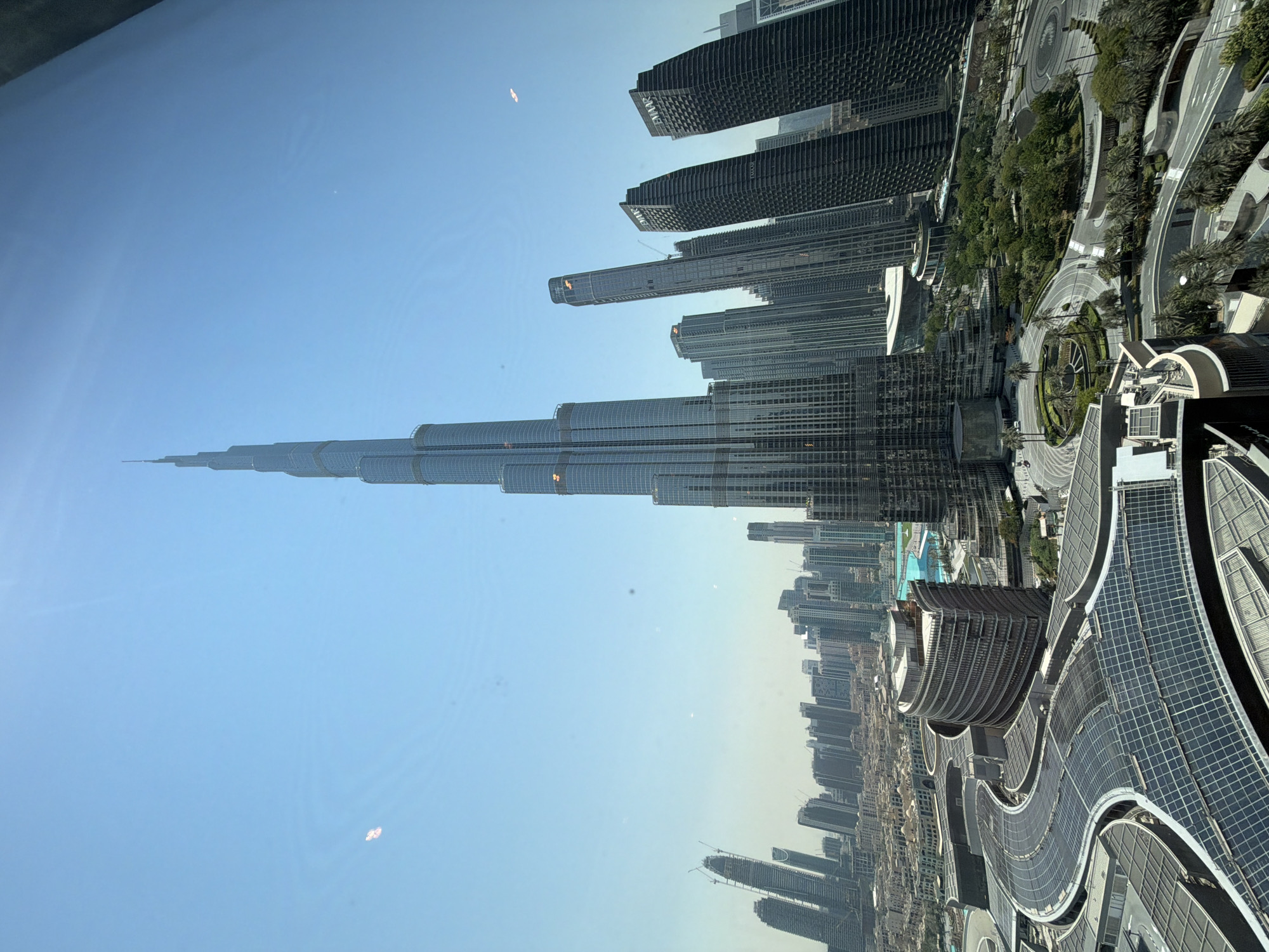 The image is a high-angle shot of the cityscape of Dubai, United Arab Emirates, dominated by the Burj Khalifa, the world's tallest building, prominently featured in the center of the frame. The sky is a clear, pale blue, with a slight haze towards the horizon.
In the foreground, an architectural complex with a distinctive curved design and vast expanses of glass roofs, likely a shopping mall or commercial area, fills the lower part of the image. These roofs are covered with solar panels, indicating a focus on renewable energy. Several other modern skyscrapers, characterized by their glass and steel facades, surround the Burj Khalifa, forming a dense urban environment. Some of these buildings are labeled with "EMAAR."
To the left of the Burj Khalifa, a body of bright blue water, likely a fountain or pool, is visible, adding a touch of color to the scene. The surrounding area appears to be a well-maintained urban landscape with roads, walkways, and green spaces featuring palm trees and other vegetation. Construction cranes are visible in the distance, suggesting ongoing development in the city. The overall impression is one of a modern, bustling metropolis with a focus on impressive architecture and urban planning.