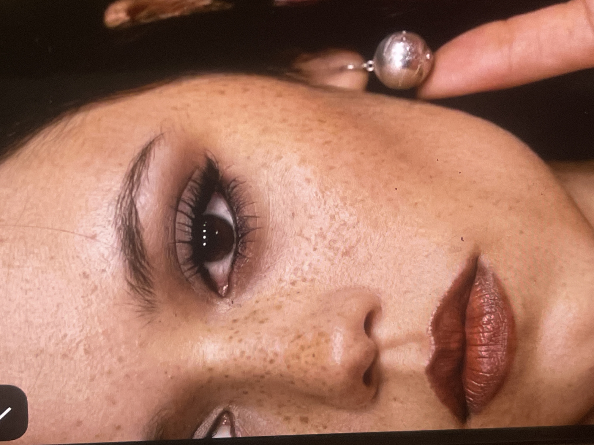 A close-up portrait photograph captures the right side of a person's face, angled upwards and to the left against a dark, out-of-focus background. The subject, likely female, has light-toned skin heavily adorned with numerous brown freckles across the cheek, nose, and forehead. Her dark eye is open, looking directly towards the viewer, with prominent, long, dark eyelashes enhanced by mascara, and a distinct eyebrow.
A well-defined nose is visible in profile, and the lips are painted a deep, textured reddish-brown color, appearing slightly parted. In the upper right portion of the frame, a light-skinned finger gently touches or holds a large, metallic, spherical earring, which reflects light and shows some surface detail. A hint of dark hair is visible along the top edge. The lighting highlights the facial features, suggesting a controlled environment like a studio. The image captures a serene or contemplative expression, focusing on beauty and detail. No time of day or specific location cues (other than the external context of Poltava, Ukraine) are discernible within the image. A white checkmark icon in a dark square is present in the bottom left corner, indicating a potential screenshot or digital overlay.