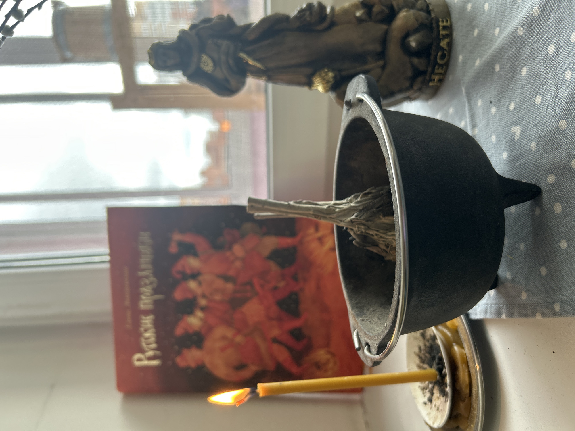 A photograph taken indoors in Balashikha, Russia, shows a small black cast iron cauldron containing dried herbs placed on a plate with a lit yellow candle. A book titled "Russian Holidays" is behind the cauldron, and a small statue of Hecate stands behind the cauldron and to the right. The background includes a window. The table or surface is covered with a blue and white polka-dot cloth.