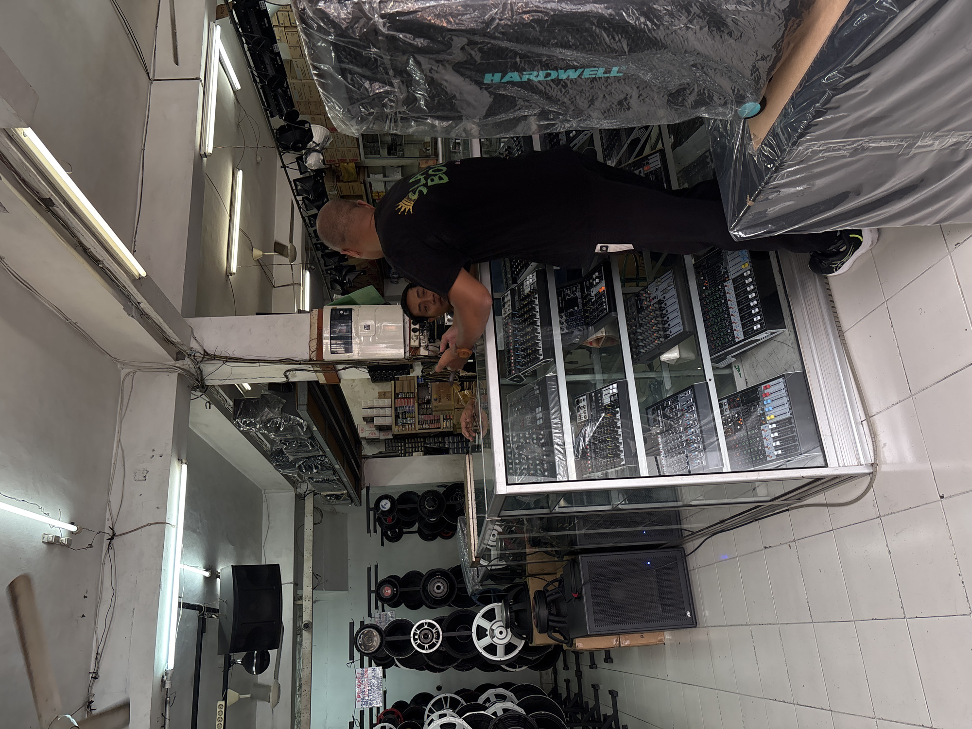 In a store located in Denpasar, Indonesia, a man with a shaved head, wearing a black t-shirt and black pants, is standing behind a glass display counter filled with audio mixers. He is interacting with another person, whose face is partially visible and appears to be looking at the man in the black shirt. The store is packed with audio equipment. On the left side of the image, numerous speaker cones are displayed vertically on racks. In the background, shelves are stocked with boxes, and other audio equipment, including speakers and lighting fixtures, are visible throughout the space. The overall environment suggests a retail establishment specializing in sound and audio systems.