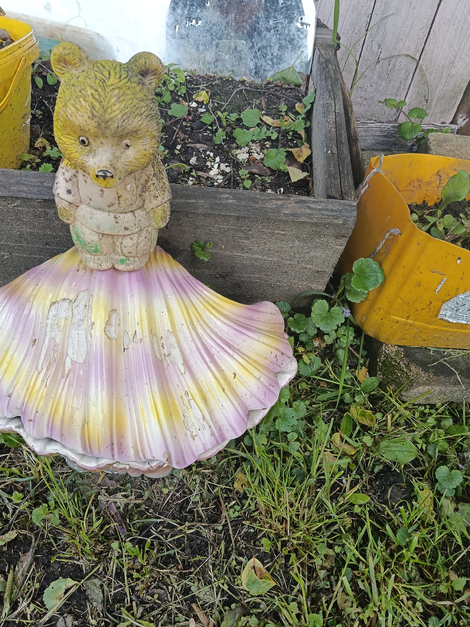 The image presents an outdoor scene in Красный Маяк, Russia, featuring a weathered, yellow teddy bear statue wearing a pink and yellow scalloped dress. The statue stands on grass and adjacent to a wooden border, possibly part of a garden bed. To the left, a yellow bucket is partially visible. To the right of the statue, a portion of a broken yellow container sits among the grass and ground cover. The scene is cluttered with grass, various small plants, and soil. The statue's coloration is faded and appears worn, suggesting that it has been exposed to the elements for an extended period.