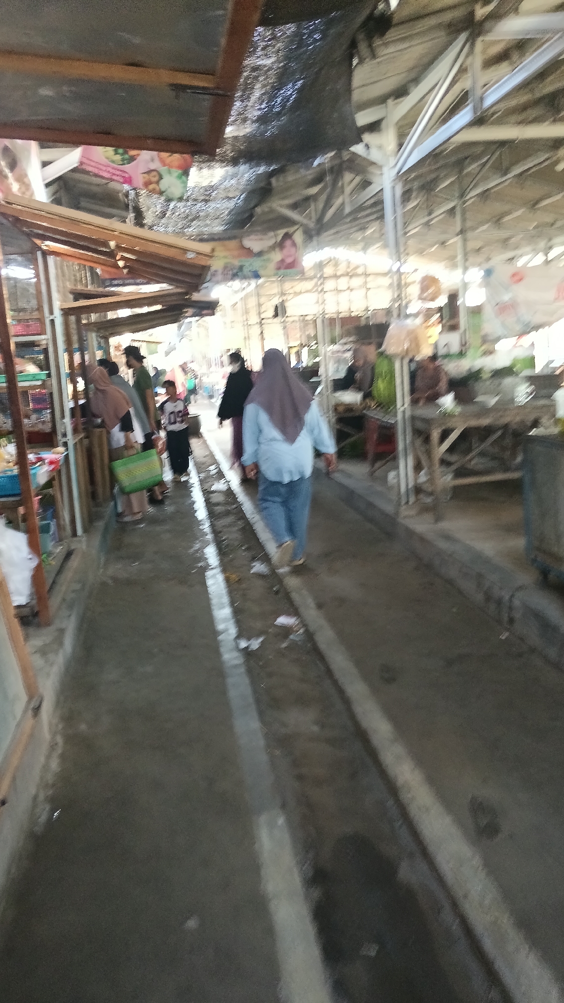 The image depicts an outdoor marketplace scene in Dukuh, Indonesia. In the foreground, a person wearing a blue long-sleeved shirt and blue pants, with a light purple hijab, walks away from the camera down a concrete pathway. The pathway appears wet and has a raised curb or gutter on either side, with some debris scattered on the ground. To the left of the pathway, a series of market stalls are visible. One stall has a green woven bag hanging from it. Several other individuals, some wearing hijabs, are seen browsing or walking within the stalls. In the background, the market structure is characterized by a metal roof supported by beams. Various signs and banners are attached to the stalls and structure. The overall environment suggests a bustling, open-air market on a day with some moisture present on the ground.