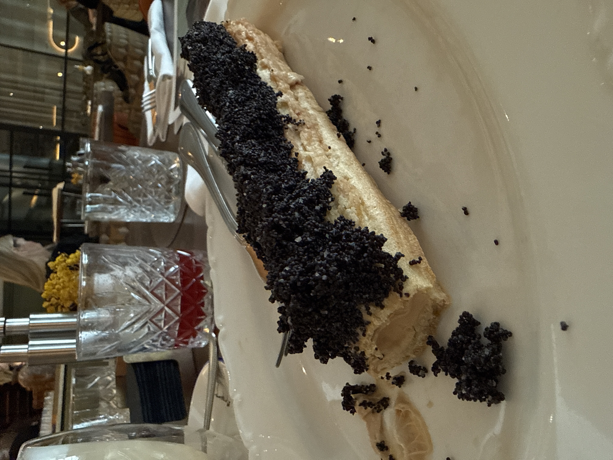 A close-up shot shows a long, light-colored pastry covered in what appears to be black poppy seeds, lying on a white plate. Cutlery is visible next to the pastry. In the blurred background are decorative glasses and a flower arrangement. The setting appears to be an indoor dining environment, likely a restaurant or cafe. While Krasnoyarsk, Russia, is not directly discernible from the image, the presence of an elaborate pastry, tableware, and glassware suggests a setting that could be found in various urban environments, including Krasnoyarsk. The details, while potentially present, are insufficient to confirm this location.