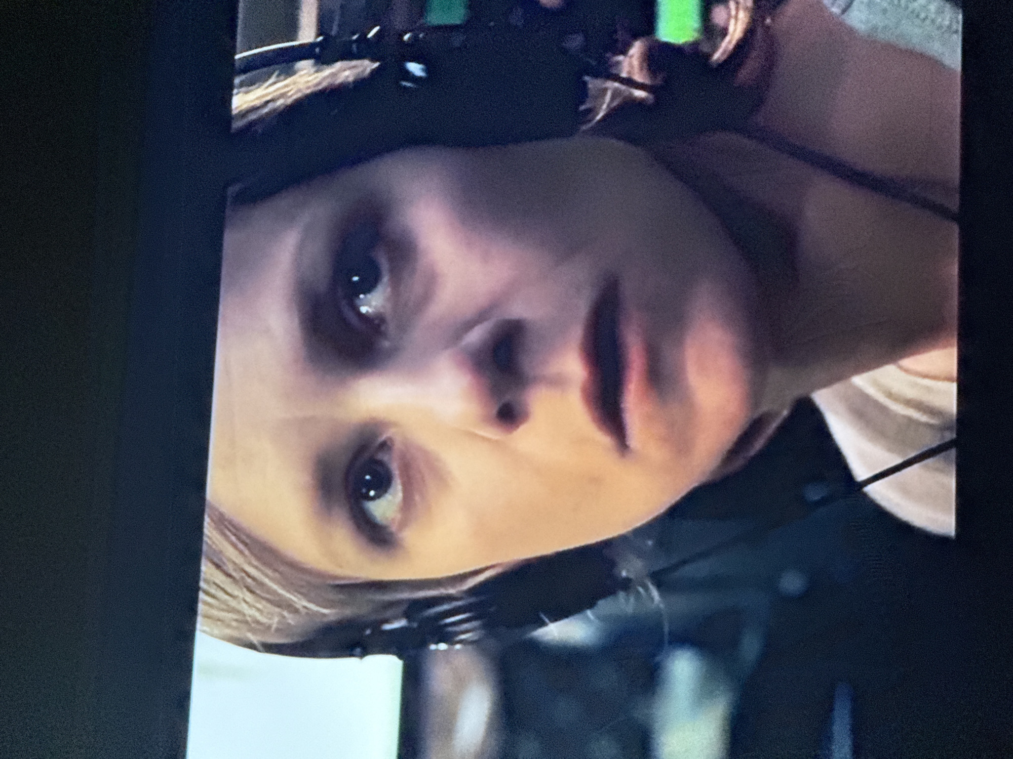 A close-up portrait depicts a fair-skinned individual with light-colored hair. The person wears a headset and is positioned at an angle, with their face occupying a significant portion of the frame. The background is blurred and indistinct. The photograph was taken on a television screen, so there is no location data available.