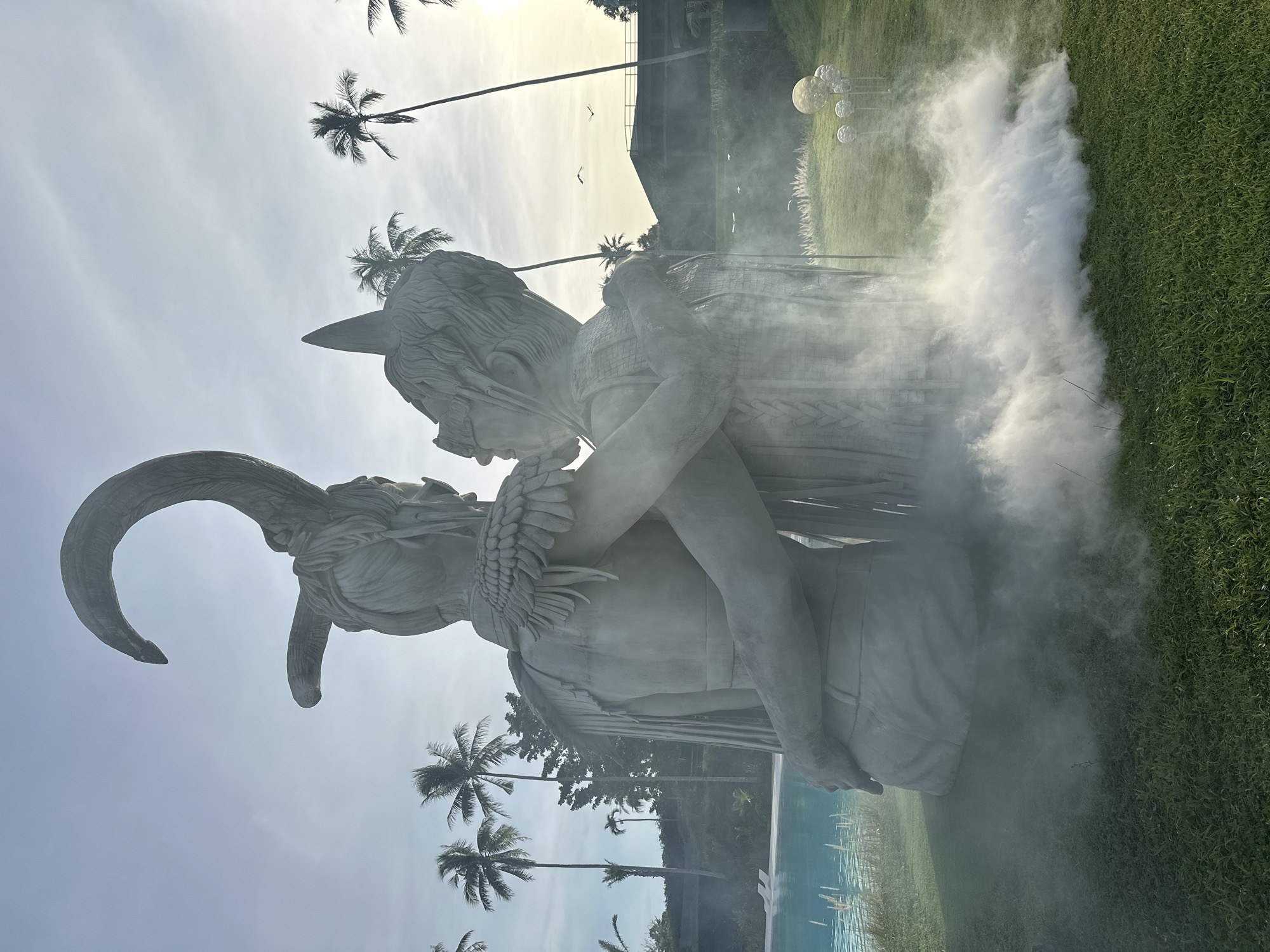 This is a vertical, eye-level, medium shot of two large concrete sculptures in an outdoor setting with a pool and grassy area. The sculptures appear to depict two figures embracing, possibly in a mythological or artistic representation. One sculpture has large horns curling downwards and backwards, while the other has a more human-like profile with feathered adornments around its neck and shoulders. The figures are rendered in a detailed, stylized manner, suggesting a contemporary art installation.
The scene is illuminated by what appears to be daylight, with the sun low in the sky, casting a soft glow. There is a noticeable haze or fog around the sculptures, particularly on the right side of the frame, obscuring some details and adding a dramatic effect. In the background, there are several tall palm trees, characteristic of a tropical location. Beyond the trees, there is a modern building structure. The ground is covered in lush green grass and a bright blue swimming pool is visible in the lower portion of the image.
The overall mood is somewhat mysterious and ethereal due to the fog and the abstract nature of the sculptures. The embracing figures and the tropical setting evoke a sense of serenity or perhaps a dramatic narrative. There is no visible text in the image. The location context of Baan Lipa Noi, Thailand, reinforces the presence of palm trees and the likely warm, humid climate.