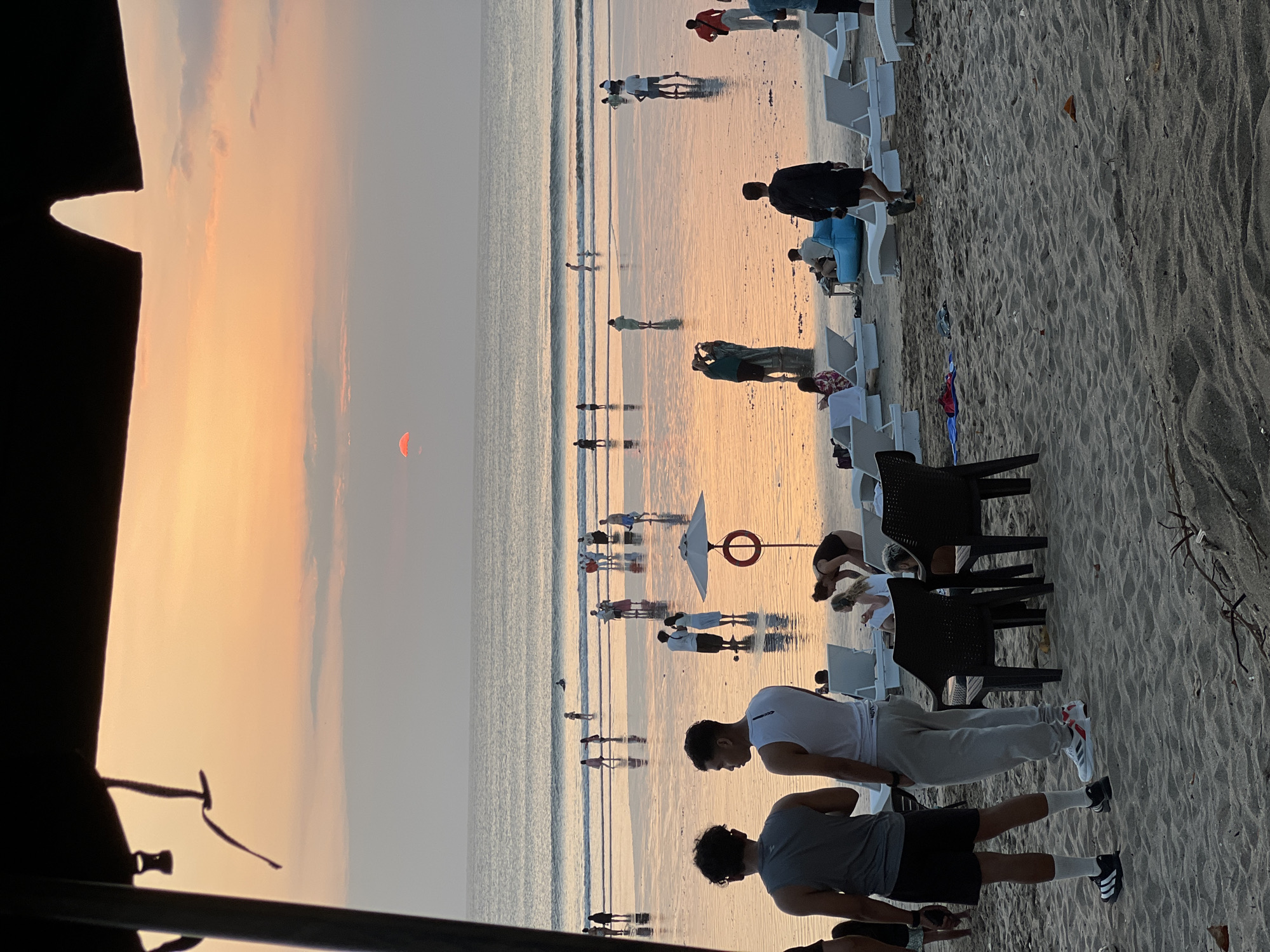 The image depicts a sunset scene on Kuta Beach, Bali, Indonesia. The sun, a vibrant orange orb, is low on the horizon, casting a warm glow across the sky and reflecting on the wet sand. The beach is populated by numerous individuals, some wading in the shallow water, others walking along the shore, and some relaxing on lounge chairs. In the foreground, two young men are standing, with the left one wearing a grey tank top and black shorts and the right one wearing a white tank top and grey sweatpants. Behind them, several dark chairs are arranged on the sand. A red life preserver ring is attached to a pole with a white umbrella nearby. The ocean in the background has gentle waves, and the sky transitions from a soft orange near the sun to a pale grey. The overall atmosphere is serene and summery, typical of a popular tourist destination like Kuta.