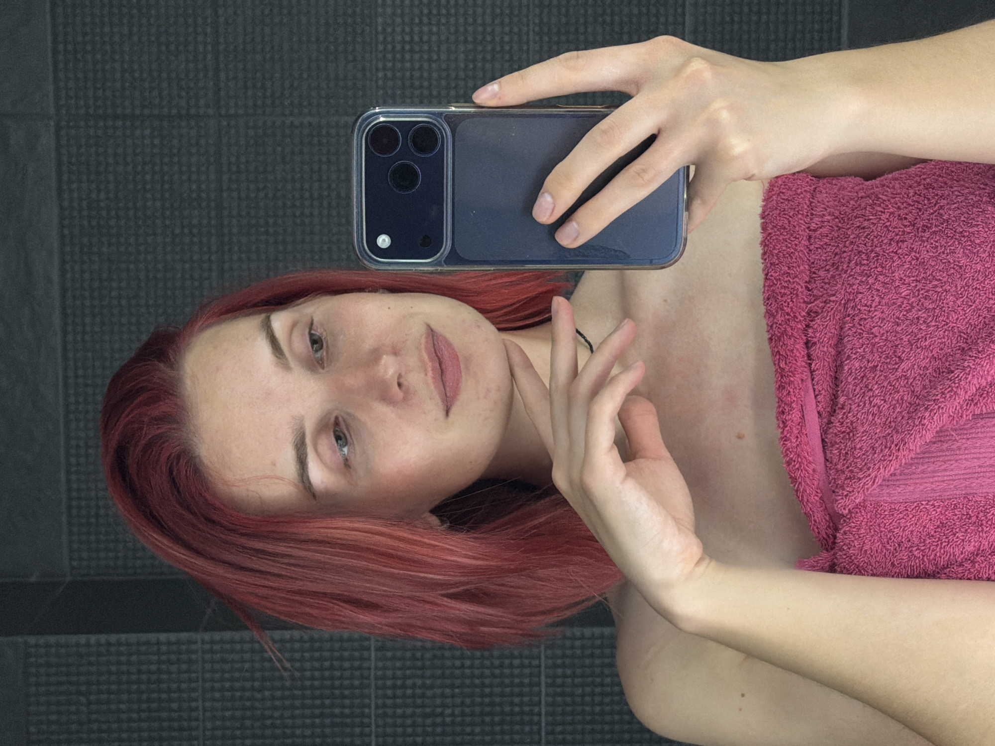 In an indoor setting, possibly a bathroom, a young Caucasian woman with short, red-dyed hair takes a mirror selfie. She is wearing a bright pink towel wrapped around her body. She is holding a dark blue smartphone in her right hand, angled to capture her reflection. Her left hand is resting gently on her chin. The background appears to be a tiled wall with a dark gray, textured finish. There are no discernable city landmarks or specific environmental indicators to suggest a location within Chaikovsky, Russia. The lighting is soft and diffuse.