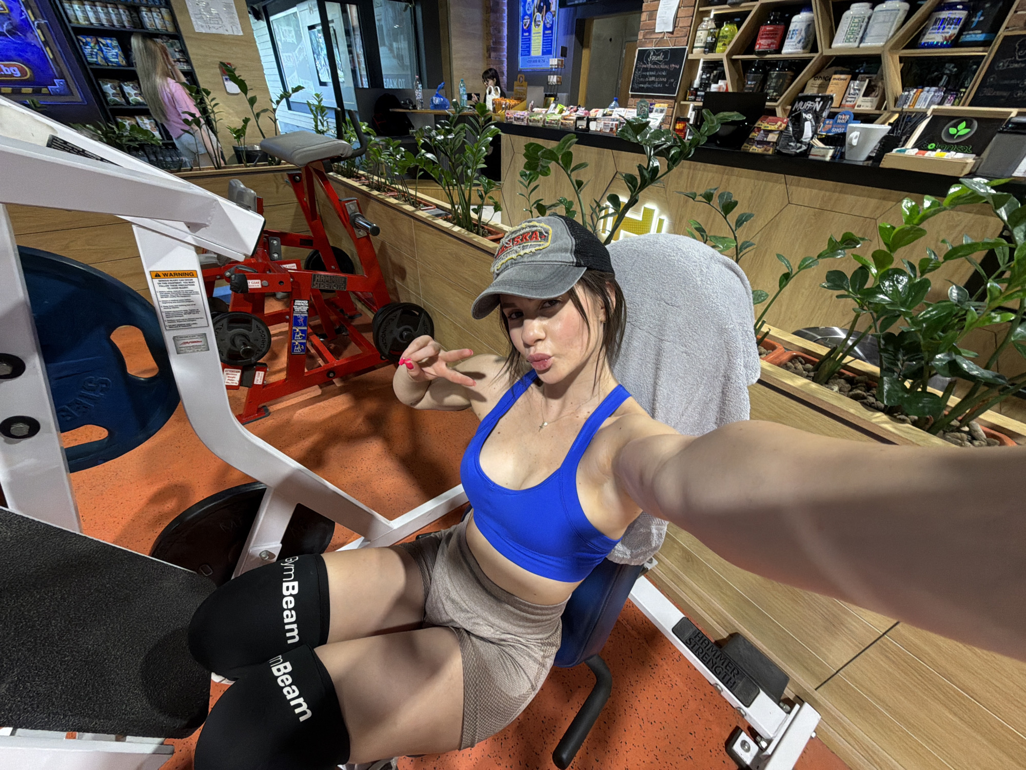 Here's a detailed description of the image:
**Scene:** The image is a selfie taken in what appears to be a gym in Plovdiv, Bulgaria.
**People:** The main subject is a young woman wearing a blue sports bra, tan exercise shorts, a gray trucker hat with the word "Alaska" on it, and black knee braces with the word "GymBeam" in white letters. She is seated on a leg press machine and making a peace sign with her left hand while pouting her lips.
In the background, partially visible, is a blonde woman wearing a pink shirt.
**Objects:** The gym equipment includes a white and black leg press machine with visible weight plates. There's also a red Hammer Strength machine in the background. Shelves with sports nutrition products, a cash register, and plants are visible along a counter area.
**Environment:** The gym has an orange and brown floor. The counter area has a faux-wood paneling. There is an open entrance to the gym.