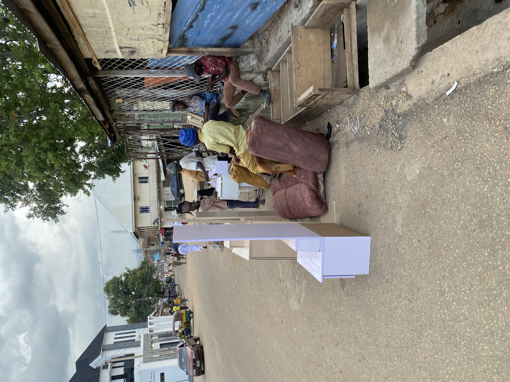 This image depicts an outdoor scene in Jos, Nigeria, likely on a street or market area. Several people are present, engaged in what appears to be a transaction or the delivery of goods. One person, wearing a red and black patterned shirt and a grey cap, is bent over and seems to be interacting with objects on the ground. Another person, in a blue and white checkered shirt and grey cap, is also visible.
The central focus of the image includes several large, cushioned items, possibly furniture like sofas or large beanbags, stacked on the ground. There is also a white, modern-looking counter or display unit positioned on the street. The setting appears to be a somewhat cluttered environment with various structures, including what looks like a makeshift stall or shop with a corrugated metal roof and blue sheeting. Wooden crates or shelving units are also visible.
In the background, more people, a tricycle (likely a "Keke Napep"), and cars are visible, suggesting a bustling urban environment. Buildings of varying architectural styles are present, including a modern white building with dark accents. The sky is overcast with clouds, indicating a potentially cloudy or pre-rainy weather condition. The overall impression is one of daily commerce and activity in a West African setting. No legible text is clearly visible in the image.