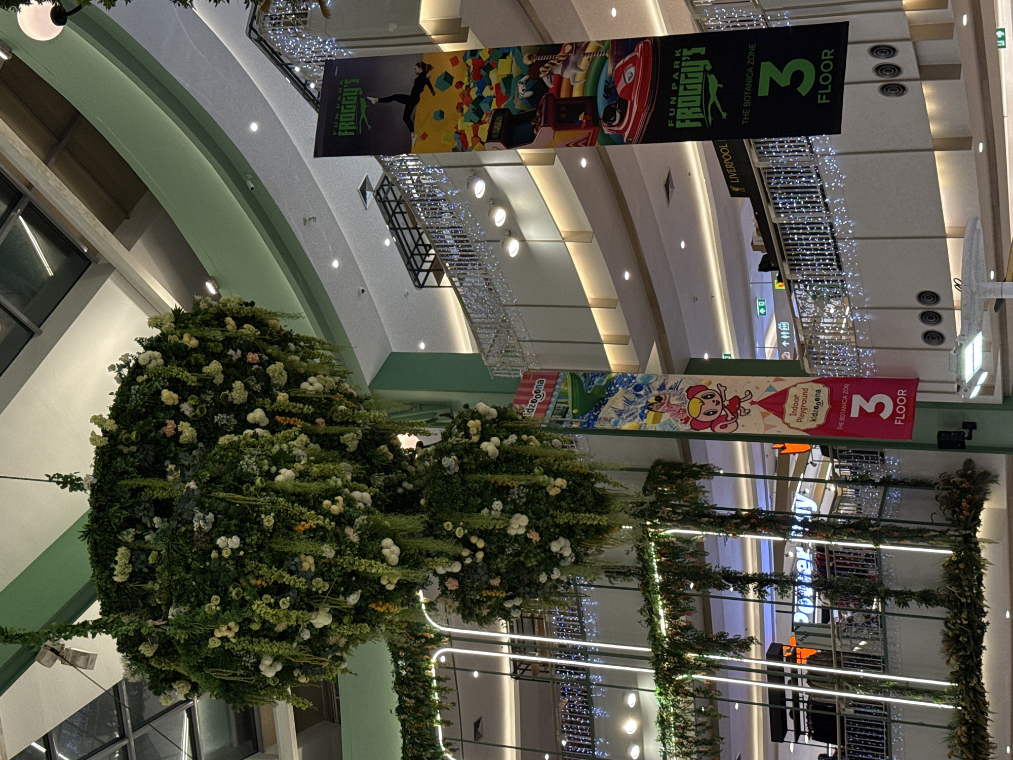This indoor scene captures the interior of a large shopping mall, likely in the city of Patong, Thailand. The focal point is the festive decorations, which include banners advertising “Froggy’s Fun Park” and “Kidzooona Indoor Playground,” along with signage indicating the location is on the 3rd floor of "The Botanica Zone" near "Liverpool." Elaborate lighting, including chandeliers and string lights, illuminates the scene. Large floral arrangements, structured into spherical shapes, and garlanded structures are prominent decorations. The overall impression is one of a commercial space decorated for a festive occasion.