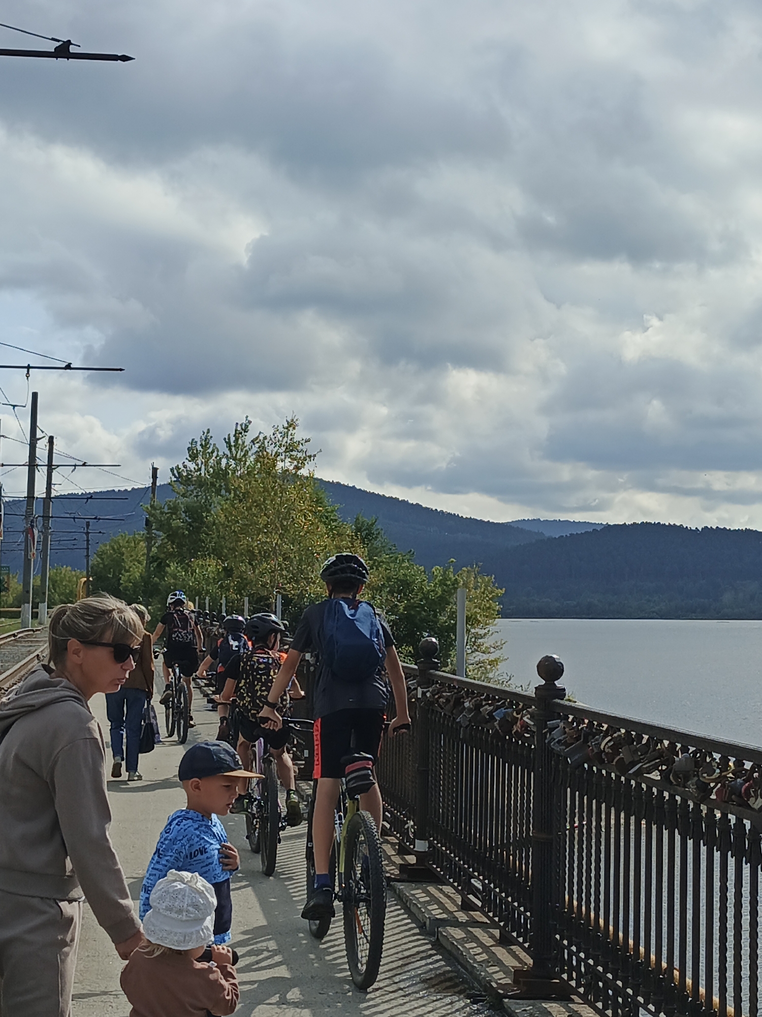 The image depicts a group of people, primarily children, on bicycles on a bridge or elevated walkway next to a body of water. The sky is cloudy. A woman in sunglasses is in the foreground on the left, and a young boy in a blue hooded sweatshirt is beside her. A very young child in a white hat is also in the foreground, facing away from the camera and holding onto something. Behind them, a procession of children on bikes, wearing helmets, proceeds along the walkway. The walkway is bordered by a decorative metal fence with many locks attached to it. In the background, there are trees, hills, and power lines. The presence of the power lines, the style of the bridge, and the mountainous terrain suggest a location like the embankment of the Ay River in Zlatoust, Russia, where similar infrastructure and natural features are found.