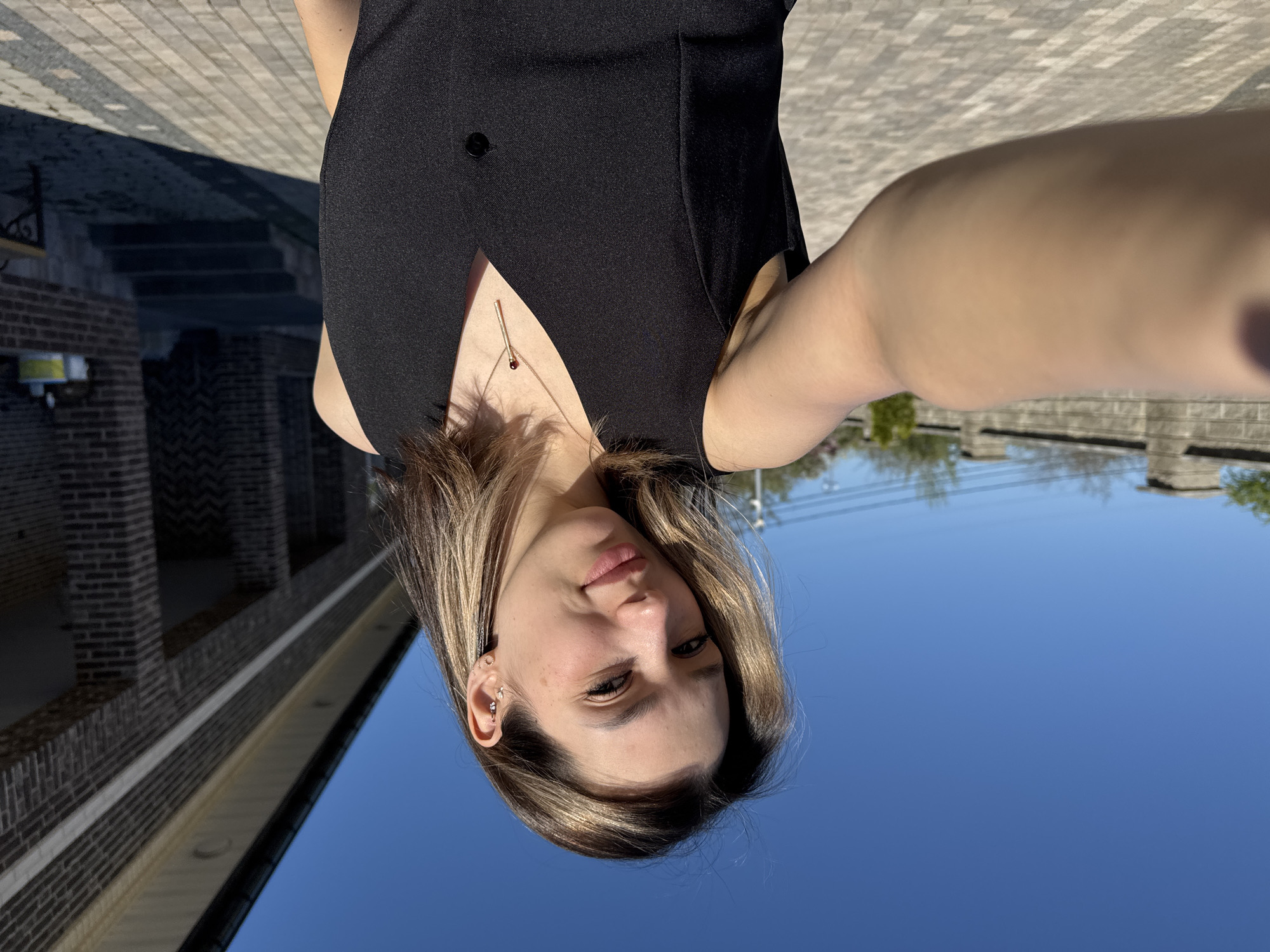 A woman stands outside, taking a selfie. She wears a black sleeveless top with a button near the neckline and a gold necklace. The background reveals an outdoor urban setting, with brick buildings and paved surfaces visible. The clear blue sky suggests a sunny day. The location is Unknown, Unknown.