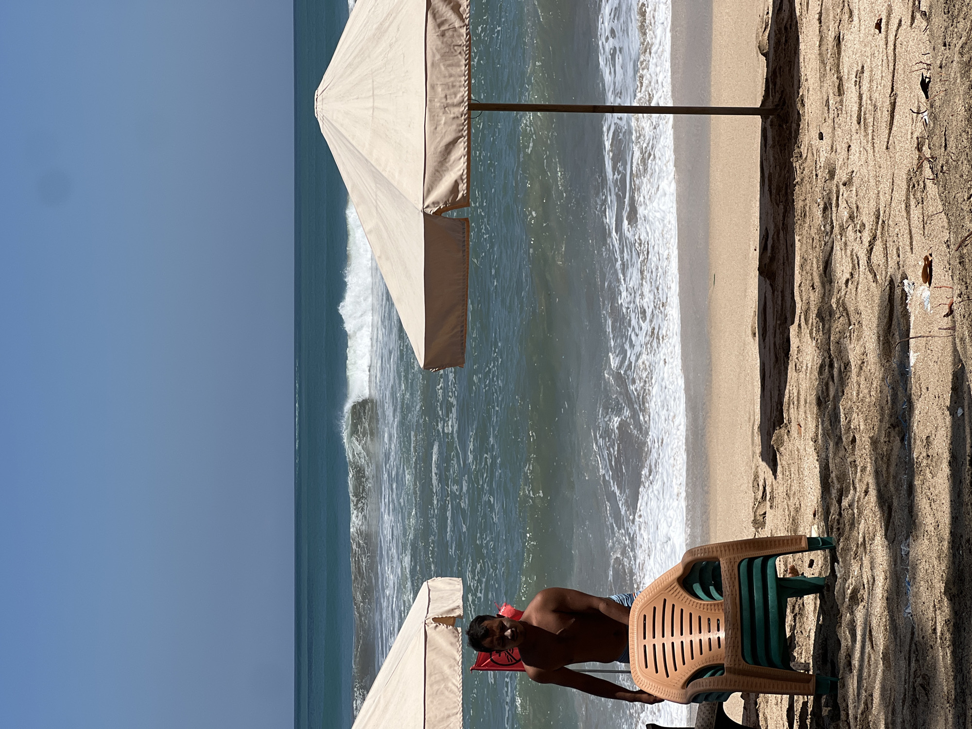 A man stands on a beach in Kuta, Indonesia, facing the ocean. He is shirtless, wearing blue shorts. In front of him are stacked plastic chairs, a beach umbrella providing shade, and a red flag with white lettering, possibly a warning sign. The ocean has moderate waves and the sky is clear and blue. The sand on the beach is light brown and scattered with small debris.