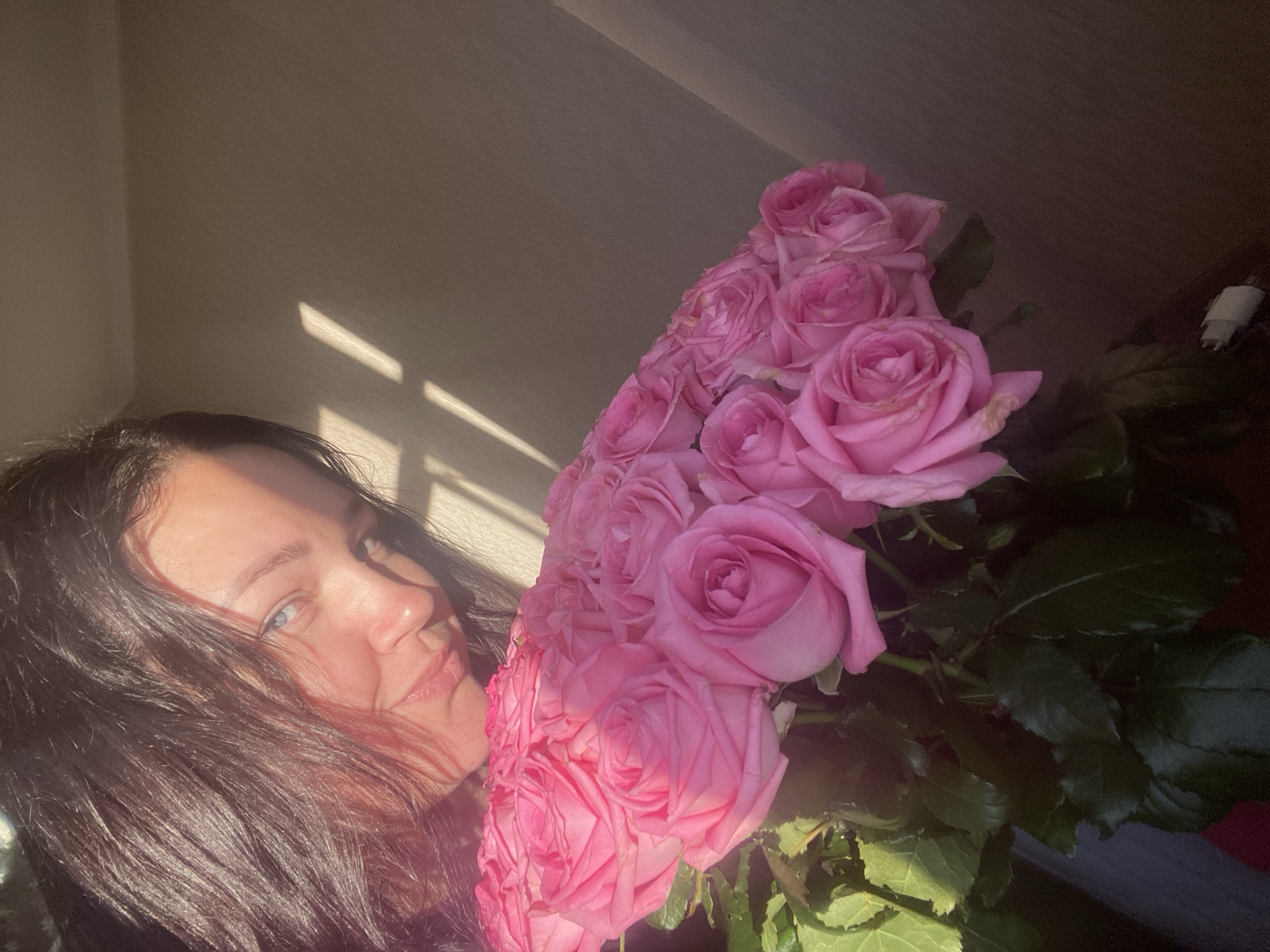 A woman with dark hair and blue eyes is smiling and holding a large bouquet of pink roses. Sunlight is casting shadows on the wall behind her. The setting appears to be indoors. There's no information to conclude it is Sochi, Russia.