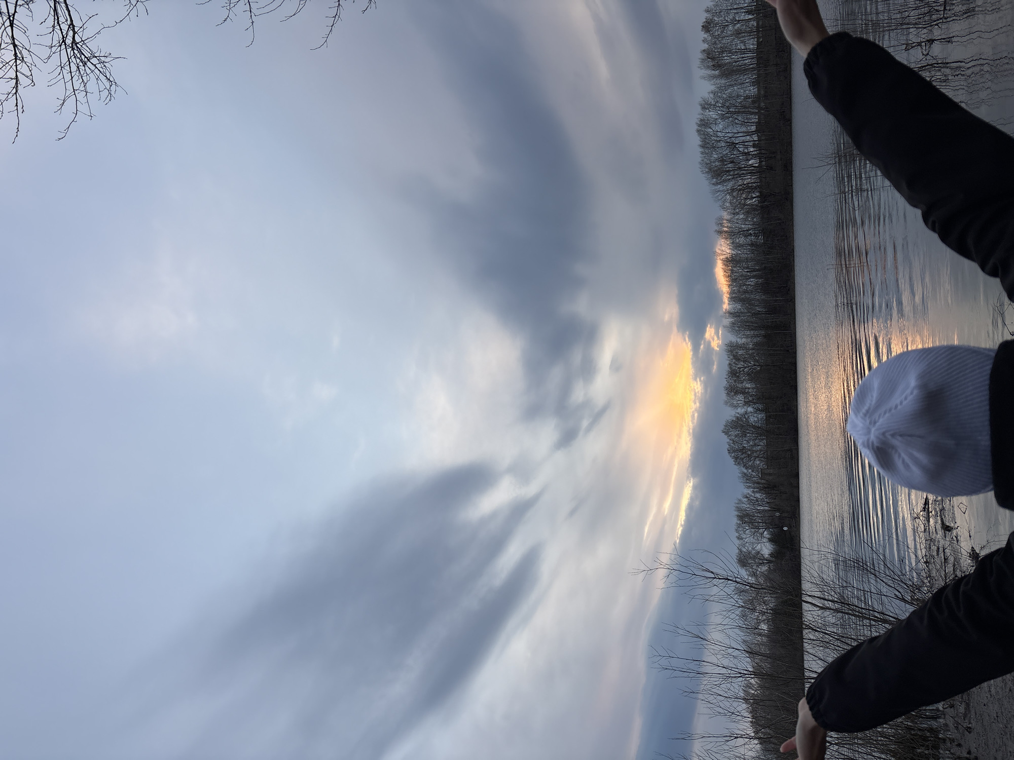The image, viewed in landscape orientation, presents a tranquil outdoor scene during either sunrise or sunset over a body of water in Chaikovsky, Russia. The sky occupies the upper two-thirds of the frame, filled with a dynamic arrangement of clouds. Predominantly, dark gray, streaky clouds stretch across the sky, contrasting with pockets of lighter, softer clouds. A striking band of bright orange and yellow light, indicative of the sun's position, cuts through the clouds on the right horizon, casting a warm glow.
Below the sky, a calm body of water, likely a lake or river, reflects the sky's colors, including the vibrant sun glow that creates a shimmering path on the water's surface. Gentle ripples are visible across the water. The distant bank is lined with a dense forest of bare trees, their dark silhouettes sharply defined against the brighter horizon, suggesting a late autumn, winter, or early spring setting.
In the bottom right corner, a person is partially visible, facing away from the viewer and observing the expansive view. They are wearing a dark jacket and a white beanie, suitable attire for cooler weather. Their arms are slightly raised, perhaps in the act of capturing the scene with a device or simply embracing the peaceful moment. The overall mood is serene and contemplative, highlighting the natural beauty of the landscape at this evocative time of day. No visible text is present in the image.