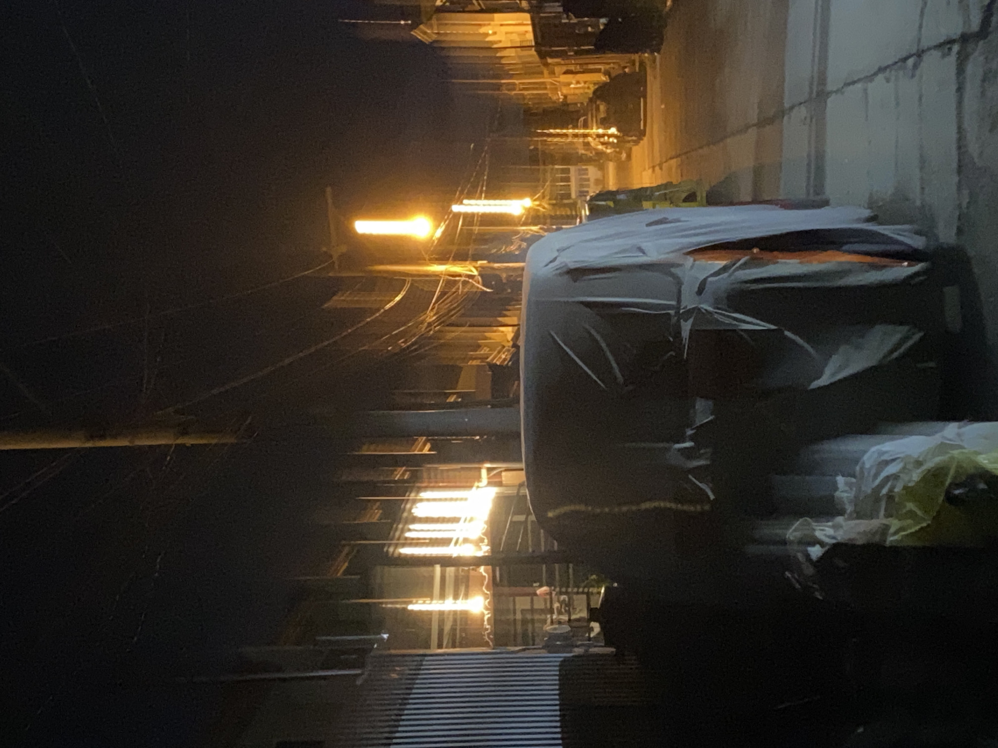 The image captures a dimly lit alleyway at night in what appears to be an urban setting, possibly in Davao City, Philippines, given the mention of a location. The scene is dominated by the muted glow of streetlights casting an orange hue on the wet pavement. In the foreground, a large object, likely a vehicle, is covered with a white tarp. Adjacent to it, black trash bags are piled up.
The alley is lined with buildings on both sides, some with what appear to be barred windows or industrial-style facades. Overhead, a complex network of power lines crisscrosses the sky, supported by wooden utility poles. Further down the alley, other vehicles are parked, their forms vaguely discernible in the low light. Some buildings exhibit illuminated windows, suggesting activity within. The overall atmosphere is one of quiet, possibly industrial, urban life after dark. There are no people visible in the frame.