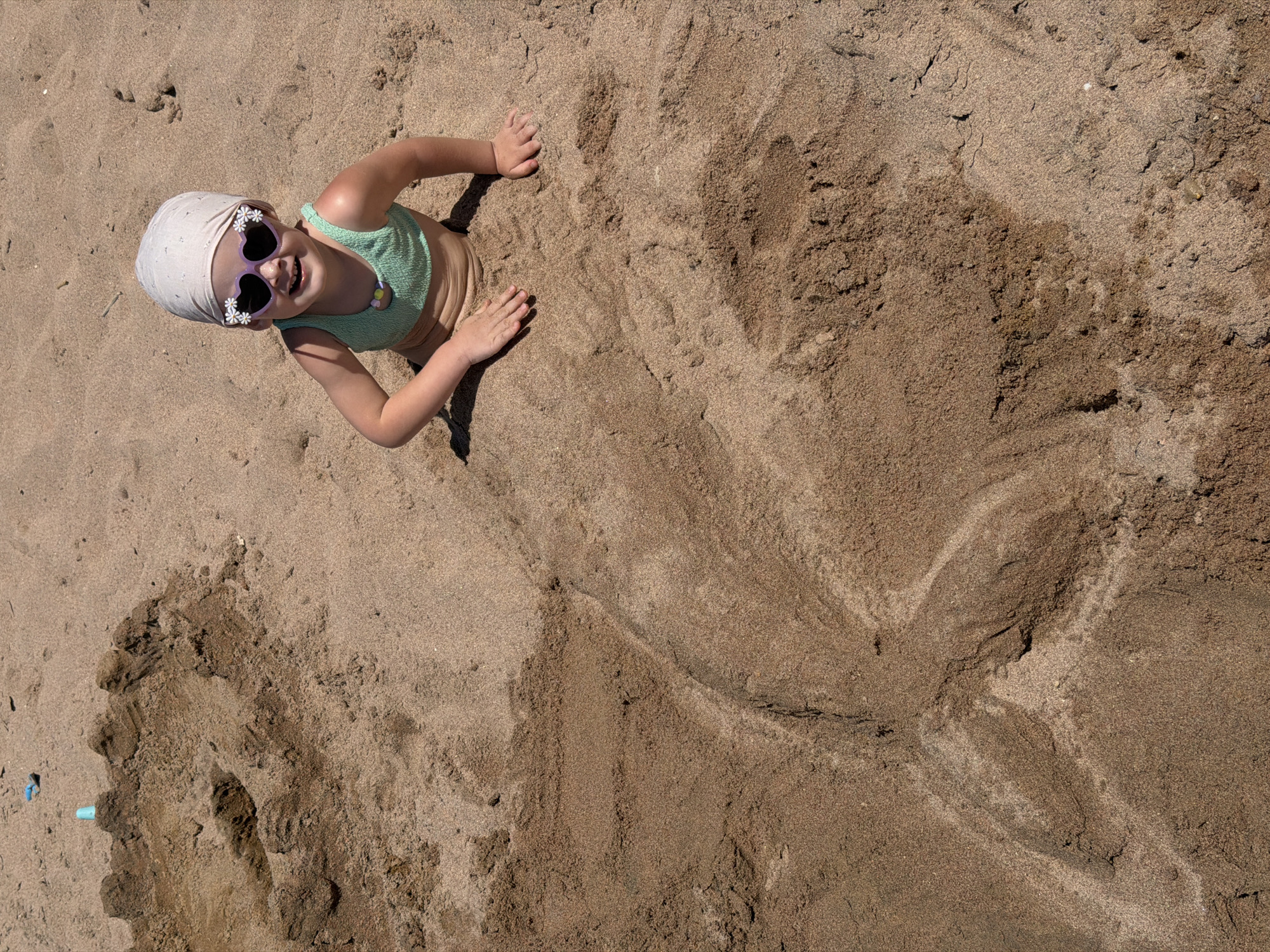 This overhead shot captures a joyful moment of a young child playing on a sunny, sandy beach. The child, likely a girl, is buried in the light brown sand from the waist down, creating the playful illusion of a mermaid's tail with sand shaped around her lower body, extending towards the right side of the frame. She is lying on her back, looking up towards the camera with a wide, beaming smile, revealing her teeth.
She is wearing a light green bikini top, a light-colored head covering, and distinctive heart-shaped sunglasses adorned with small white flowers. A colorful beaded necklace is also visible around her neck. Her arms are resting on the sand beside her torso.
The activity is clearly beach play, with the child immersed in the sand. The bright daylight creates clear shadows, indicating a sunny day. The sandy environment is consistent with a beach setting, supported by the given context of Sharm El Sheikh, Egypt. A small, light blue plastic object is partially visible in the sand in the bottom left corner. The child's happy expression and the creative sand sculpture convey a sense of fun and relaxation during a beach outing. There is no visible text in the image.
