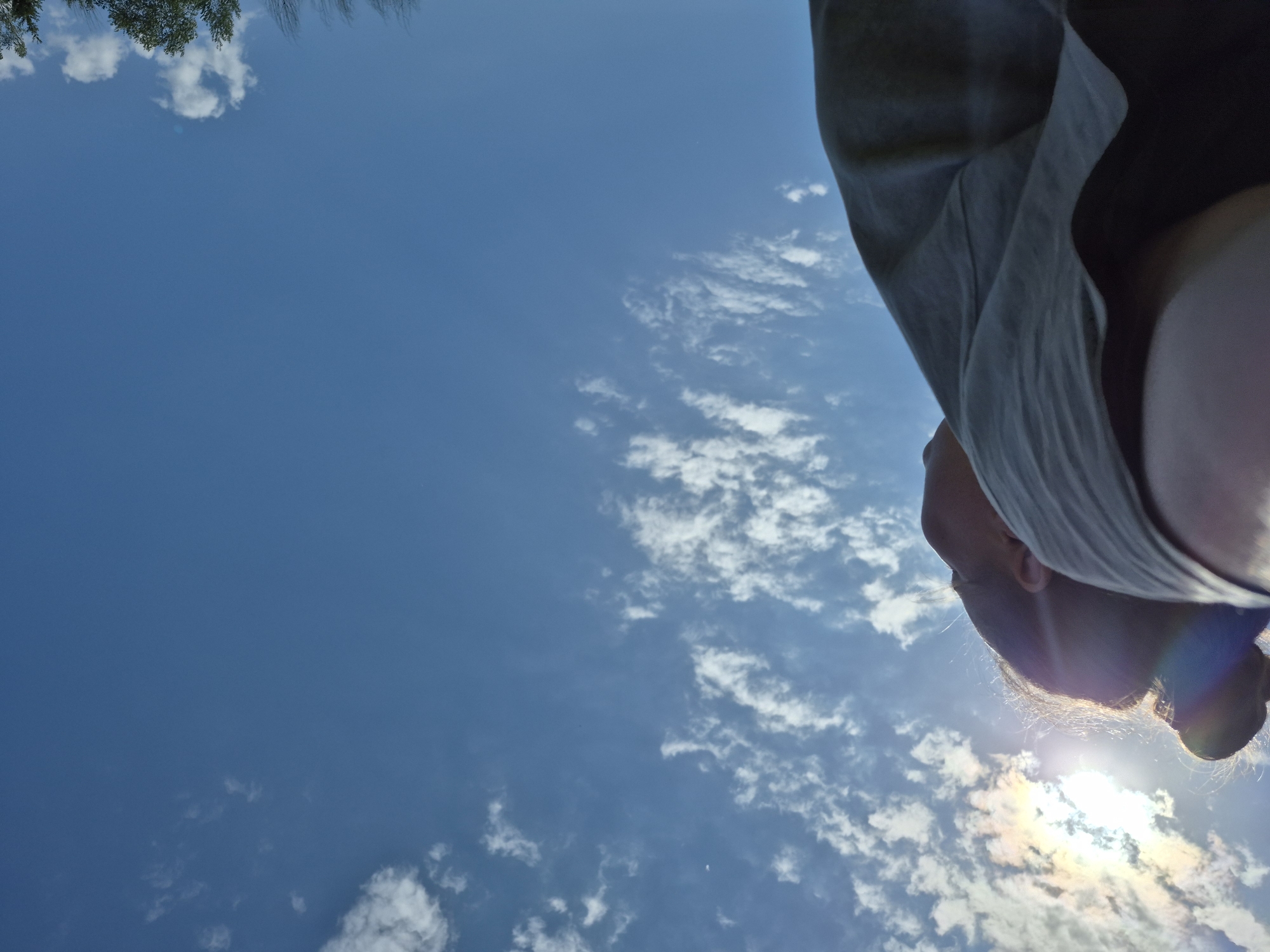 The photo is a low-angle shot looking up into a blue sky with scattered white clouds. In the lower right portion of the frame, a person's head and upper body are visible, partially obscuring the sky. The sun is behind the person's head, creating a halo effect and a rainbow-colored lens flare. The person is wearing a light gray garment. Some trees are visible in the upper left. There is no indication of the specific location being in Дойная, Russia.