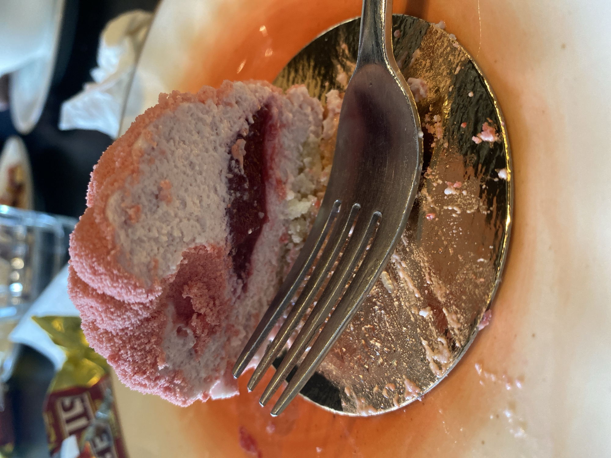 The image shows a close-up view of a partially eaten pink and purple cake on a golden circular plate. A silver fork is resting on the plate, with its tines partially inserted into the cake. The cake appears to have layers of pink frosting and a pink crumb coating. There are crumbs and pink liquid on the plate, suggesting the cake has been recently consumed. In the background, out of focus, are hints of a white napkin and a red-hued surface. A portion of a yellow and red wrapper with visible text "ST" is also visible on the left side of the image. The lighting suggests an indoor setting.