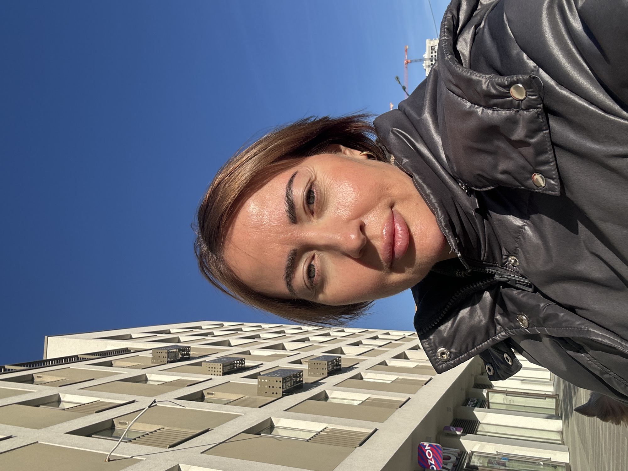 A woman in a black jacket takes a selfie from a low angle on a sunny day. Behind her is a tall, light-colored building with a grid-like pattern of windows and small structures on the roof. In the distance, construction cranes can be seen against the clear blue sky. The woman is smiling, with her face angled toward the camera. The scene appears to be in Yekaterinburg, Russia.