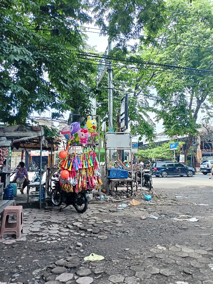 This media file captures a bustling street corner in Mojosari, Indonesia, during the daytime. The central focus is a motorcycle used as a mobile vendor's stall, heavily adorned with colorful party supplies. These include numerous bright party horns or noisemakers in various colors, some with shiny tinsel, and several foil balloons featuring popular characters like Hello Kitty and a yellow pineapple-like figure, along with some plain orange spherical balloons.
To the left, under a rustic canopy or lean-to, a person in a patterned shirt and dark pants sits on a wooden bench, next to large blue water dispenser bottles. The ground in the foreground is rough and uneven, consisting of cracked pavement and dirt, with scattered litter. Large, lush green trees frame the top of the image, providing shade over the scene, while numerous power lines crisscross overhead.
In the mid-ground, there are other simple stalls or structures. The street extends into the background where a dark SUV and other vehicles are visible, indicating a busy road. Several utility poles and signs line the street. One distant blue street sign is partially visible, and another sign on a pole near the center has some black text that is not clearly legible. Another sign further back might contain the text "Ayam Bakar." The overall scene depicts informal commerce thriving in an everyday urban environment.