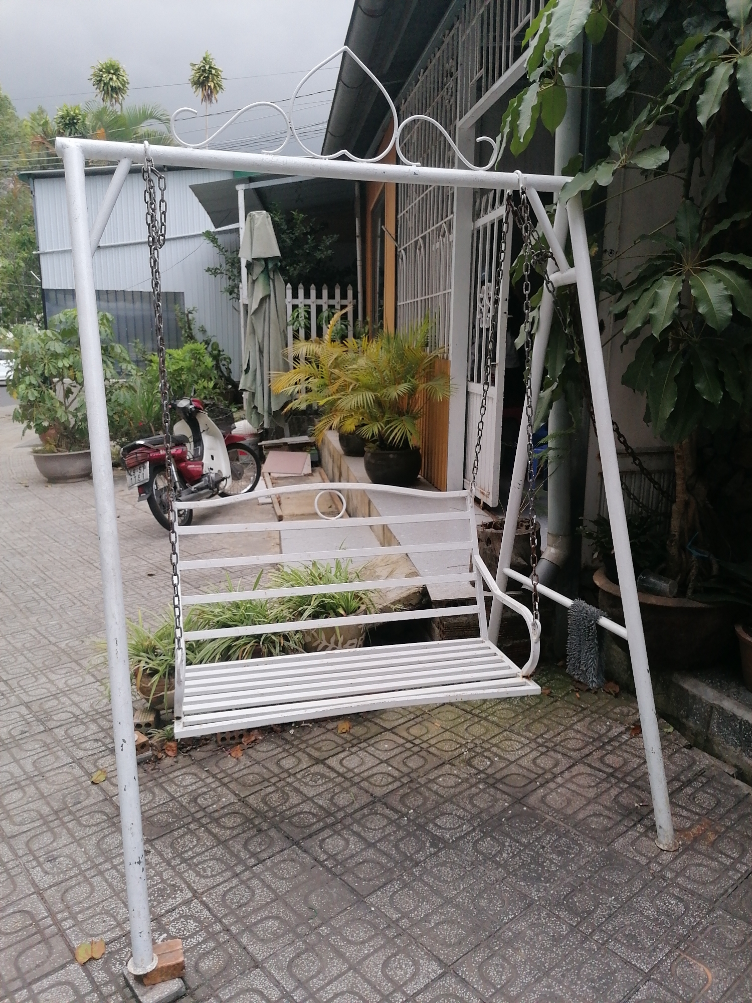 The image depicts a white metal swing set situated on a tiled patio in Da Lat, Vietnam. The swing set features a slatted seat and is suspended by chains from a white metal frame. To the left of the swing set, a red and white motorcycle is parked. Various potted plants are visible around the patio, and in the background, there's a building with a white fence and an umbrella. The sky is overcast, suggesting a cloudy day. The ground is paved with patterned tiles, and there are some scattered leaves. The overall scene appears to be an outdoor residential area or a small establishment.