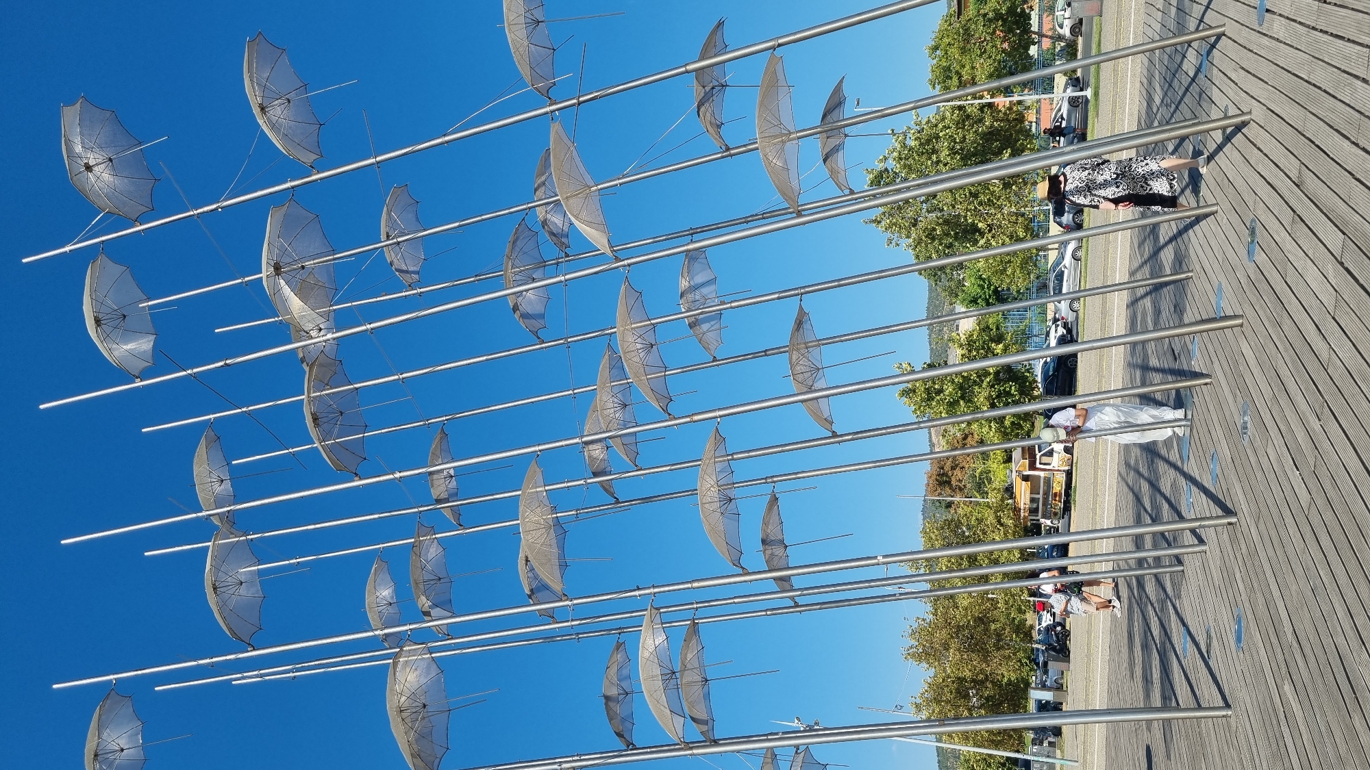 The photograph captures a section of the Umbrellas sculpture by George Zongolopoulos, located on the Thessaloniki waterfront in Greece. The sculpture features numerous metallic poles supporting transparent umbrella-like structures, all set against a clear blue sky. In the foreground, a wooden boardwalk, characteristic of the waterfront promenade, is visible, with several metallic poles casting shadows. Two individuals are present on the boardwalk: one person in a white outfit is leaning down, seemingly interacting with the base of a pole, while another person wearing a patterned dress and a hat is walking away from the viewer. In the background, trees, parked cars, a food truck, and a distant hillside are discernible, indicating an urban coastal environment.