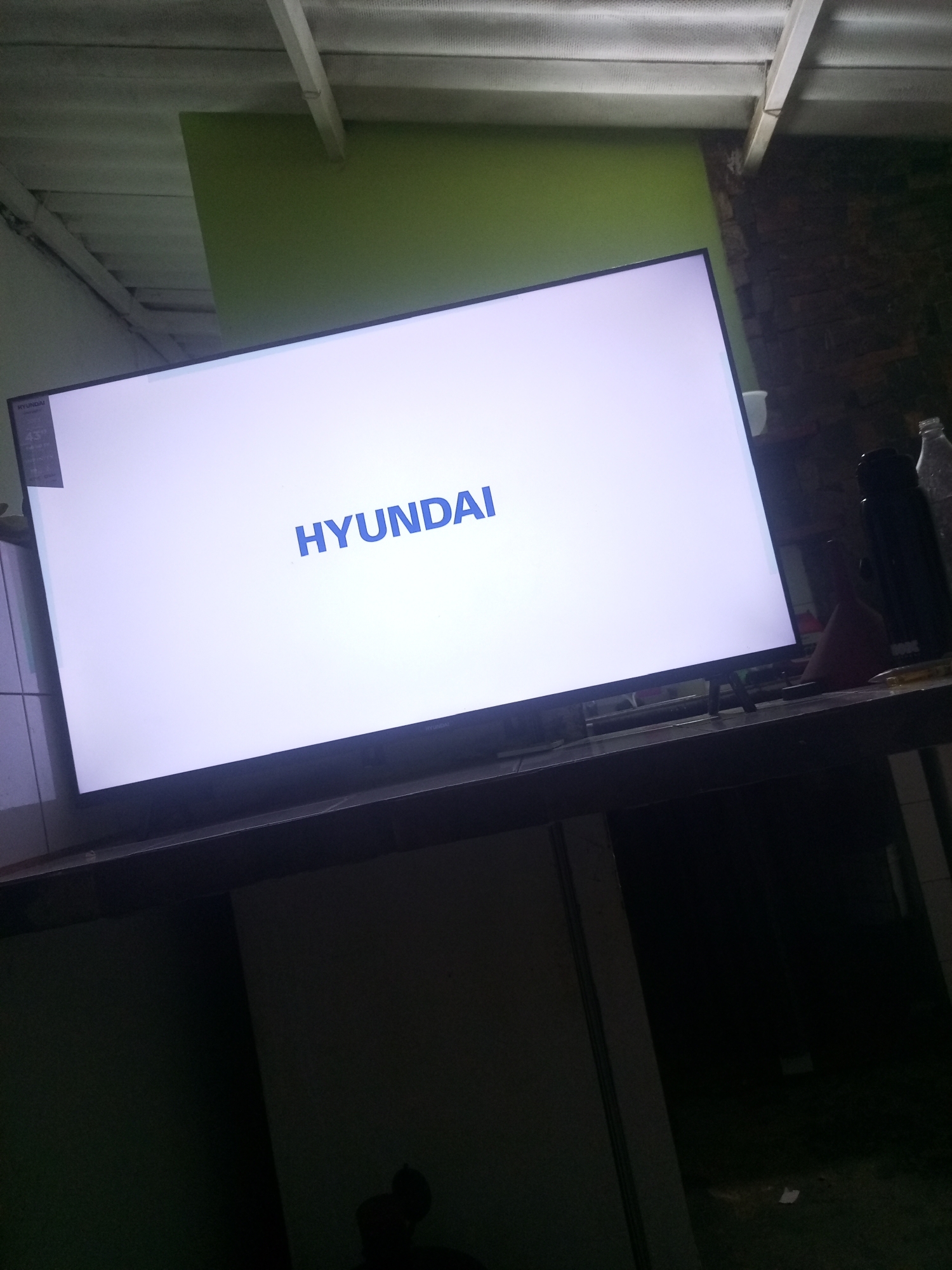 In the city of La Tagua, Venezuela, a large, white television screen displays the blue "HYUNDAI" logo. The TV is positioned on a dark, reflective surface, likely a table or counter. In the background, a rough brick wall is visible, and above, a corrugated metal ceiling with white beams is present. The lighting appears dim, with some ambient light reflecting off the screen. To the right of the TV, a black thermos and a clear plastic bottle are also present. The overall scene suggests an indoor setting, possibly a home or workshop, with the TV being the central focus.