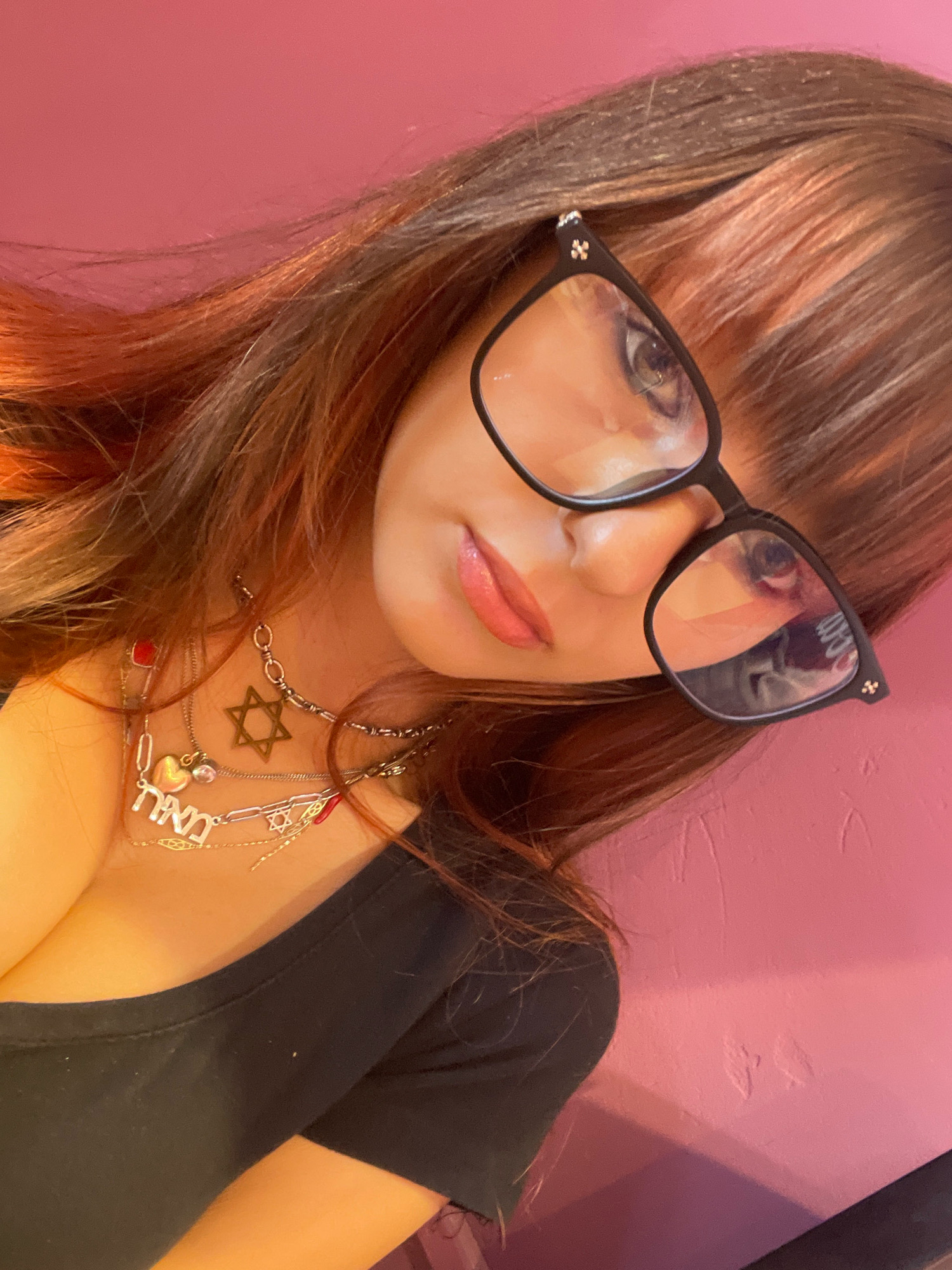 A woman with long, auburn-highlighted hair and eyeglasses is shown from the chest up, looking slightly to her left. She is wearing a black top and several necklaces, including one with a Star of David pendant and another with Hebrew letters. The background is a solid pink wall with some textured marks on it. The image appears to be a selfie taken indoors, likely in a residential setting. It is not possible to determine the specific location within Raanana, Israel.