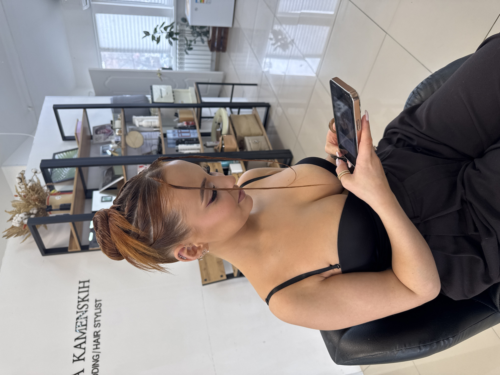 An indoor scene depicts a young adult female seated in a black leather chair within a hair salon or beauty studio in Chaikovsky, Russia. A sign on the white wall behind her reads "A KAMENSKIH WEDDING/HAIR STYLIST." The woman has reddish-brown hair styled in an updo with shaved sides and wears a black strapless top and dark trousers. Multiple piercings are visible on her left ear, and she wears rings on her right hand, which has manicured nails. She is observed looking down at and interacting with a metallic-framed smartphone held in both hands. The background features a black shelving unit stocked with beauty products, a white door, a window with horizontal blinds, a radiator, a small potted plant, and a white compact refrigerator. The floor is composed of light-colored, reflective tiles.