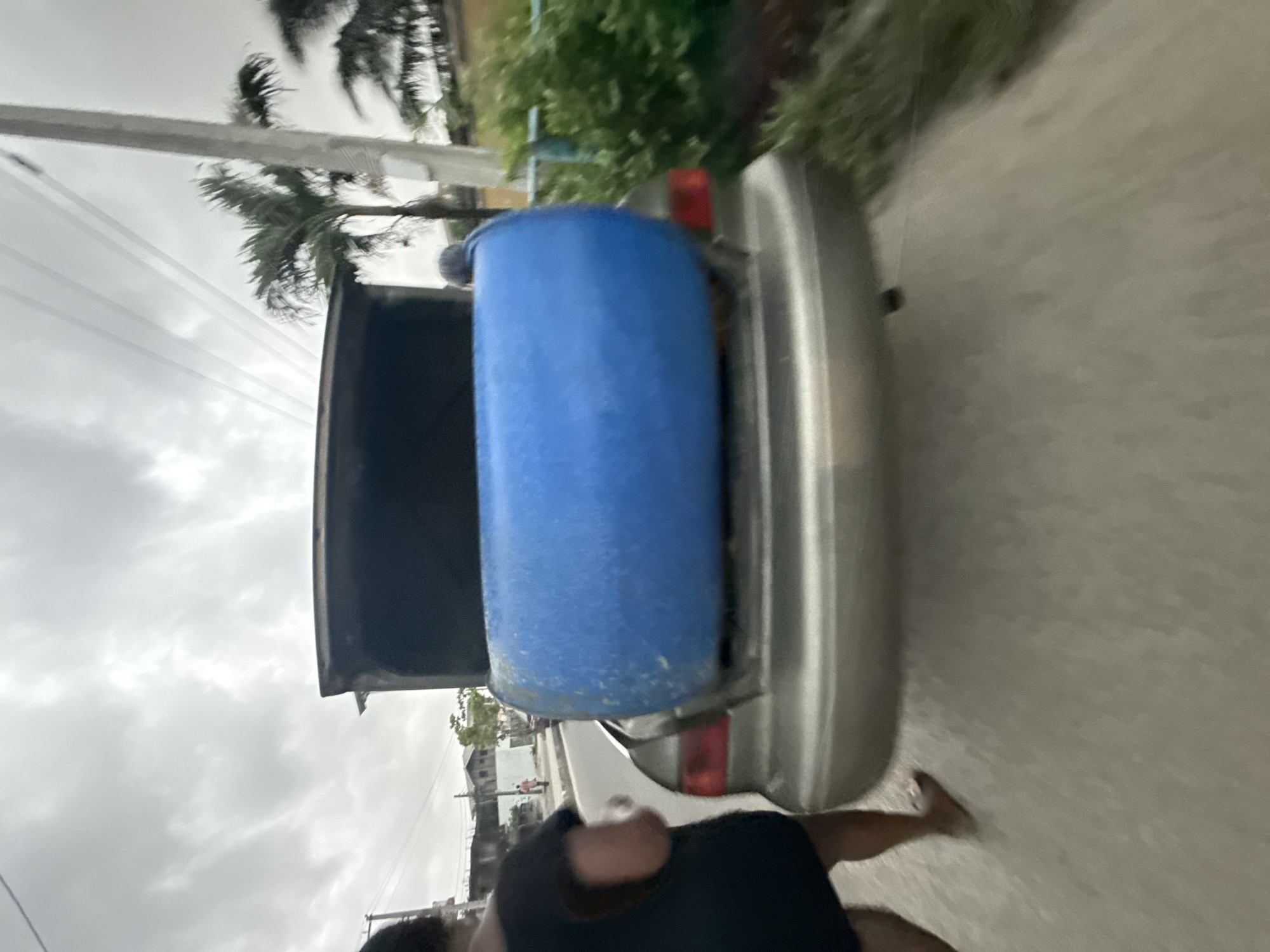 The image, originally rotated, shows an outdoor street scene in Ado, Nigeria, under an overcast sky. A silver or light-colored sedan car with its trunk open is prominent in the foreground, facing towards the right. Inside the open trunk, a large, blue plastic barrel or drum is placed horizontally, suggesting it is being transported. The car's red taillight is partially visible on the right side of the trunk.
To the immediate left of the car, a person is partially visible, wearing a dark shirt. Their arms and legs are also discernible, indicating they are either standing or walking beside the vehicle. The road surface beneath the car and person appears to be asphalt or concrete.
The background features elements typical of a tropical environment. On the left, utility poles with power lines stretch across the frame, with lush green palm trees visible behind them. On the right, green foliage, likely bushes and trees, creates a verdant backdrop, and faint outlines of buildings can be seen in the distance. The sky is predominantly cloudy, indicating an overcast daytime. There is no visible text in the image, and no specific emotions are discernible from the subjects.