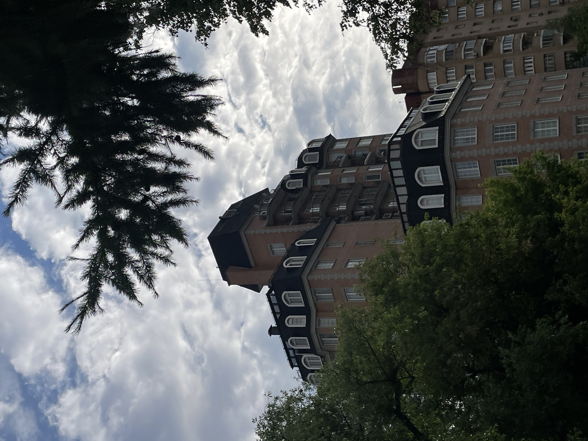 The image depicts a multi-story building with an ornate facade, possibly an apartment complex or hotel, nestled amongst lush green trees. The architecture is characterized by a mansard roof with arched dormer windows, and the brickwork is accented with decorative stone elements. The building is situated in a moderately urban environment, with hints of other structures visible in the background. The sky above is partly cloudy, with a mix of sunlight and shade, suggesting an overcast or late afternoon setting. The scene appears peaceful and serene, with the trees providing a natural contrast to the man-made structure.