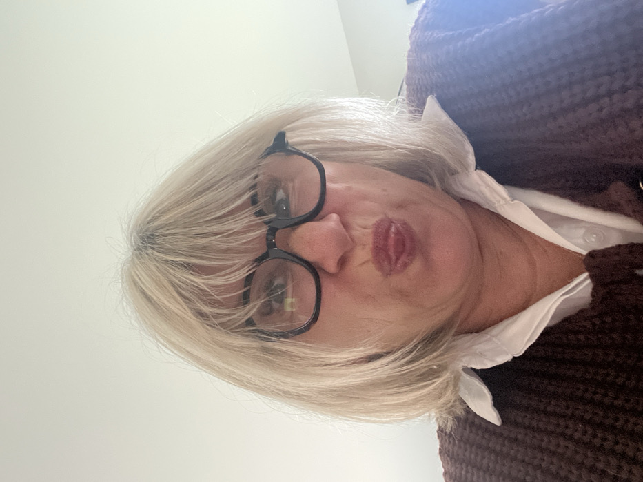 A close-up, rotated selfie shows an adult individual with short, platinum blonde hair and black-framed glasses. The image is oriented such that the person's head is tilted to the right, implying they are looking down or up at the camera. Their face is centered in the frame, capturing a playful or informal expression with pursed lips, often referred to as a "duck face" or "kissy face," and a hint of reddish-brown lipstick.
The blonde hair is styled loosely around their face, partially covering their forehead and ears. Visible roots show a slightly darker shade, possibly their natural color or gray. Their eyes are visible behind the clear lenses of the black, rectangular-framed glasses.
The person is wearing a white collared shirt, visible around the neckline, beneath a chunky, dark brown or reddish-brown knitted sweater or cardigan. This outer garment is visible primarily on the right side of the frame.
The background is uniformly light-colored, suggesting an indoor setting with a plain wall or ceiling. A bright glare or reflection is noticeable in the upper right corner of the image, indicating a light source nearby. The lighting is bright and even across the person's face. There is no visible text in the image. The overall impression is an informal, personal photograph.