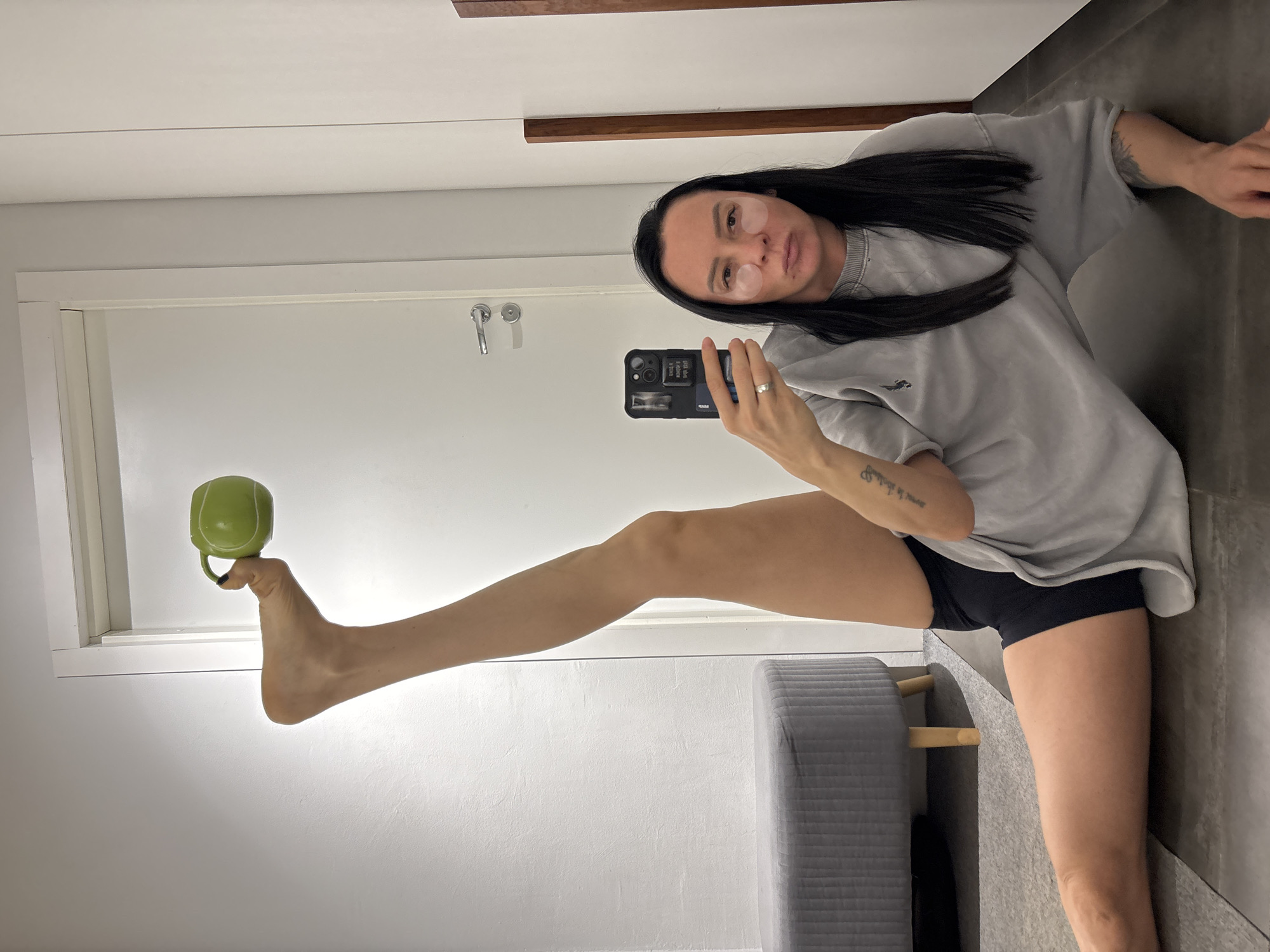 A woman is taking a selfie in what appears to be a bathroom or dressing room. She is lying down, with one leg extended upwards, holding a green mug on her foot. She is wearing a gray t-shirt, black shorts, and has eye patches on. She is holding a black smartphone in her hand, presumably taking the picture. The background includes a white door, gray flooring, and a gray ottoman. The location is not possible to determine as the image does not contain any explicit signs identifying the city of Chaikovsky, Russia.