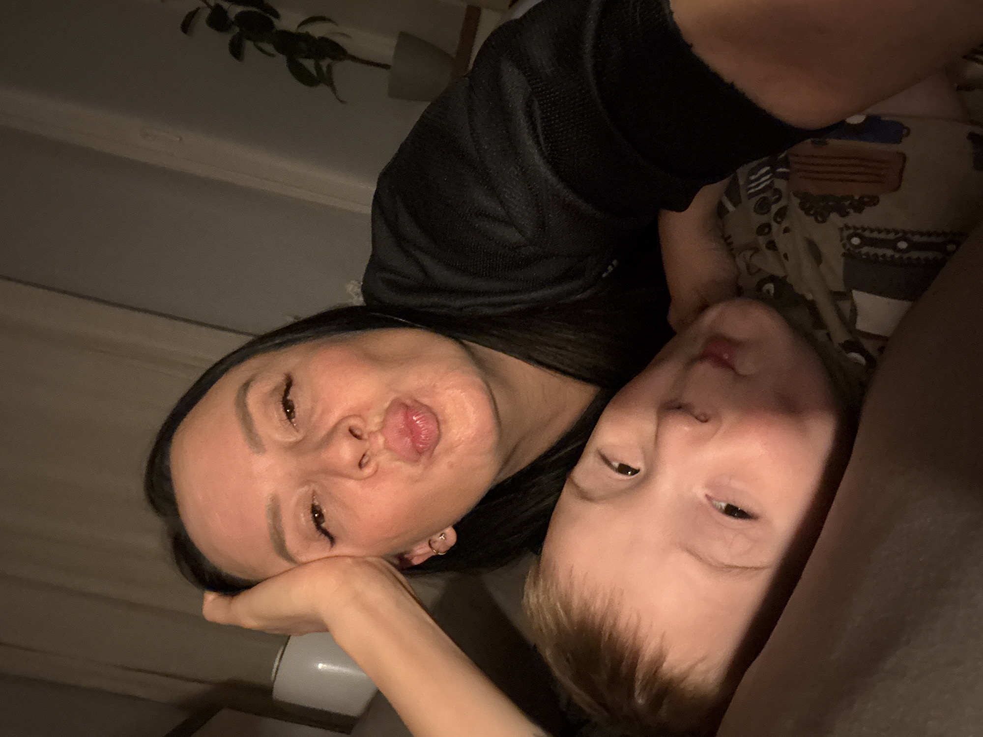 This indoor, rotated selfie-style portrait features an adult woman and a young child, likely a mother and her son, in a home in Chaikovsky, Russia. The image is taken from a high angle, looking down at both subjects who are lying close together, creating a sense of intimacy.
The woman, positioned to the left, has long dark hair and is wearing a black, textured top with a visible mesh-like pattern. Her head is resting on her left hand, which appears to be holding a white cup or mug. She is making a playful "duck face" with puckered lips, indicating a lighthearted and affectionate mood. A small, circular earring is visible on her left ear.
To the right of the woman is the child, who has light brown hair and dark eyes. The child looks towards the camera with a calm or slightly sleepy expression. The child is wearing a patterned garment, possibly pajamas, featuring various shapes and motifs in muted colors.
The setting appears to be a dimly lit indoor space, suggesting it is evening or night. In the upper left background, a dark green leafy plant is visible against a light-colored wall or ceiling. The soft, low lighting contributes to a cozy and relaxed atmosphere. There is no visible text in the image. The scene captures a tender and playful moment between the two individuals.