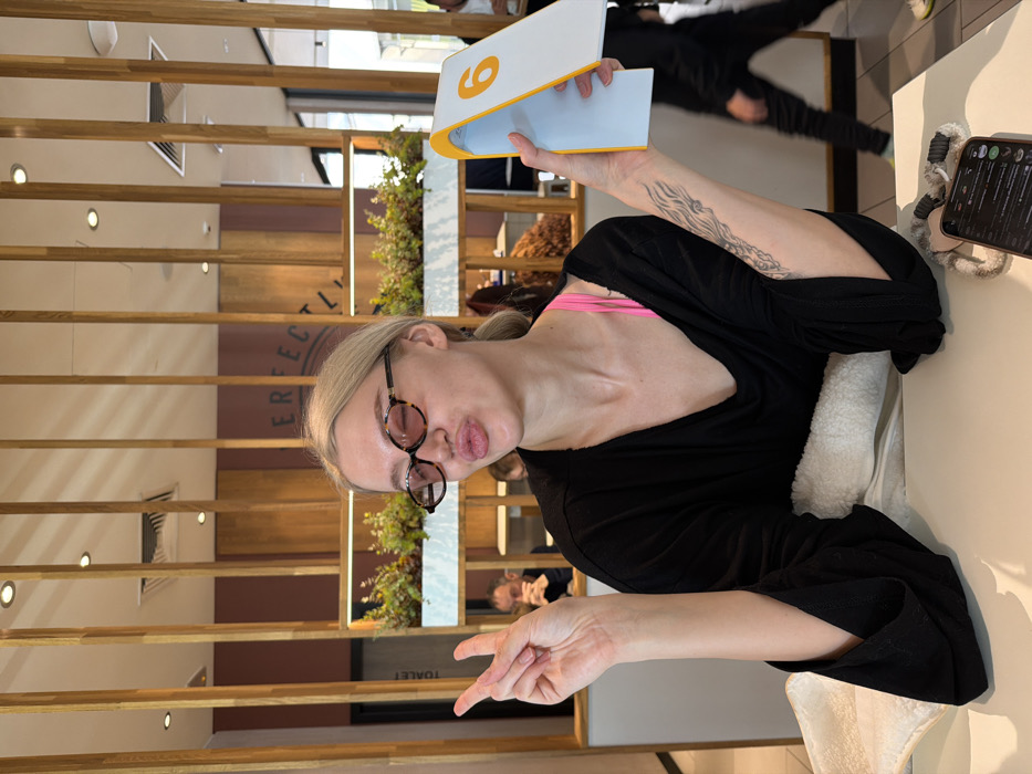 A young blonde-haired female, estimated to be in her twenties, is seated at a light-colored table within a modern cafe or restaurant. She wears tortoiseshell-patterned oval sunglasses with pink lenses and a loose-fitting black v-neck top, revealing a bright pink bra strap. Her left hand is raised, displaying a peace sign gesture. Her right hand holds a white and yellow table number stand, prominently displaying the number "6." A black tattoo featuring a flowing, abstract design is visible on her right forearm. Her lips are pursed in a pout. A smartphone displaying a dark interface, along with a grey and white coiled accessory, rests on the table to her right.
The background features decorative vertical wooden slats and horizontal wooden panels. Behind the subject, a white planter box contains green foliage. A partial sign reading "PERFECTL" is visible on a light brown wall. To the far left, a door is marked with the Serbian word "TOALET." Blurred figures of other patrons are visible throughout the background, including a man behind the subject's left shoulder and another person walking in the far right distance. The overall setting is consistent with a contemporary dining establishment.
The location is identified as Perfectly Food & Bar at Kneza Miloša 92, Belgrade, Serbia, based on the visible branding ("PERFECTL" for "Perfectly") and the "TOALET" sign indicating a Serbian-speaking environment.