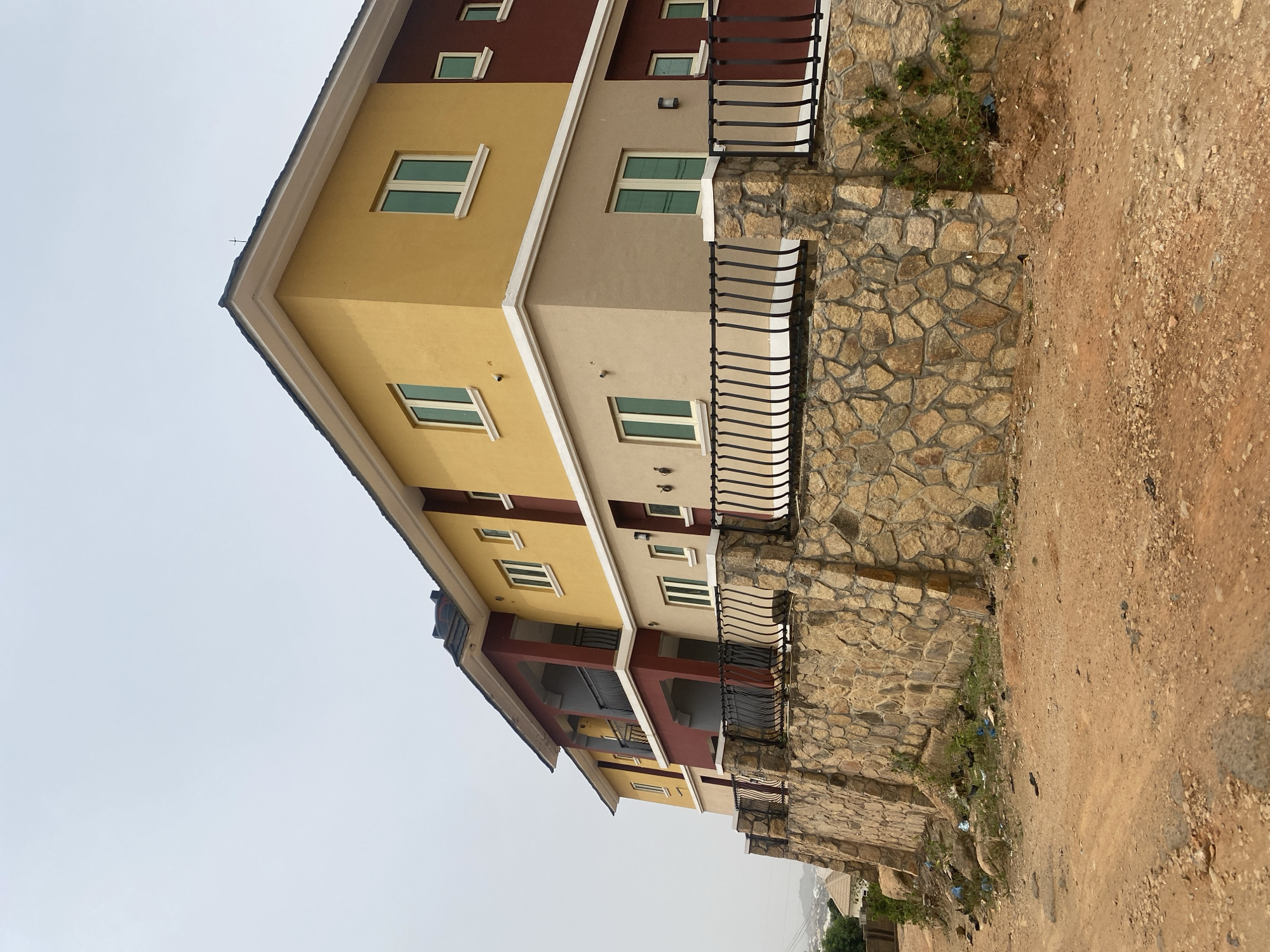 The image shows a three-story building located in Barakin Akawo, Nigeria. The building's exterior is painted in alternating sections of yellow, tan, and brown. The windows are rectangular with white frames. A black metal railing runs along the front of the building, situated atop a wall constructed of irregular stones. The ground in front of the building is dirt, with sparse vegetation and scattered trash. The sky is overcast. No people are visible.