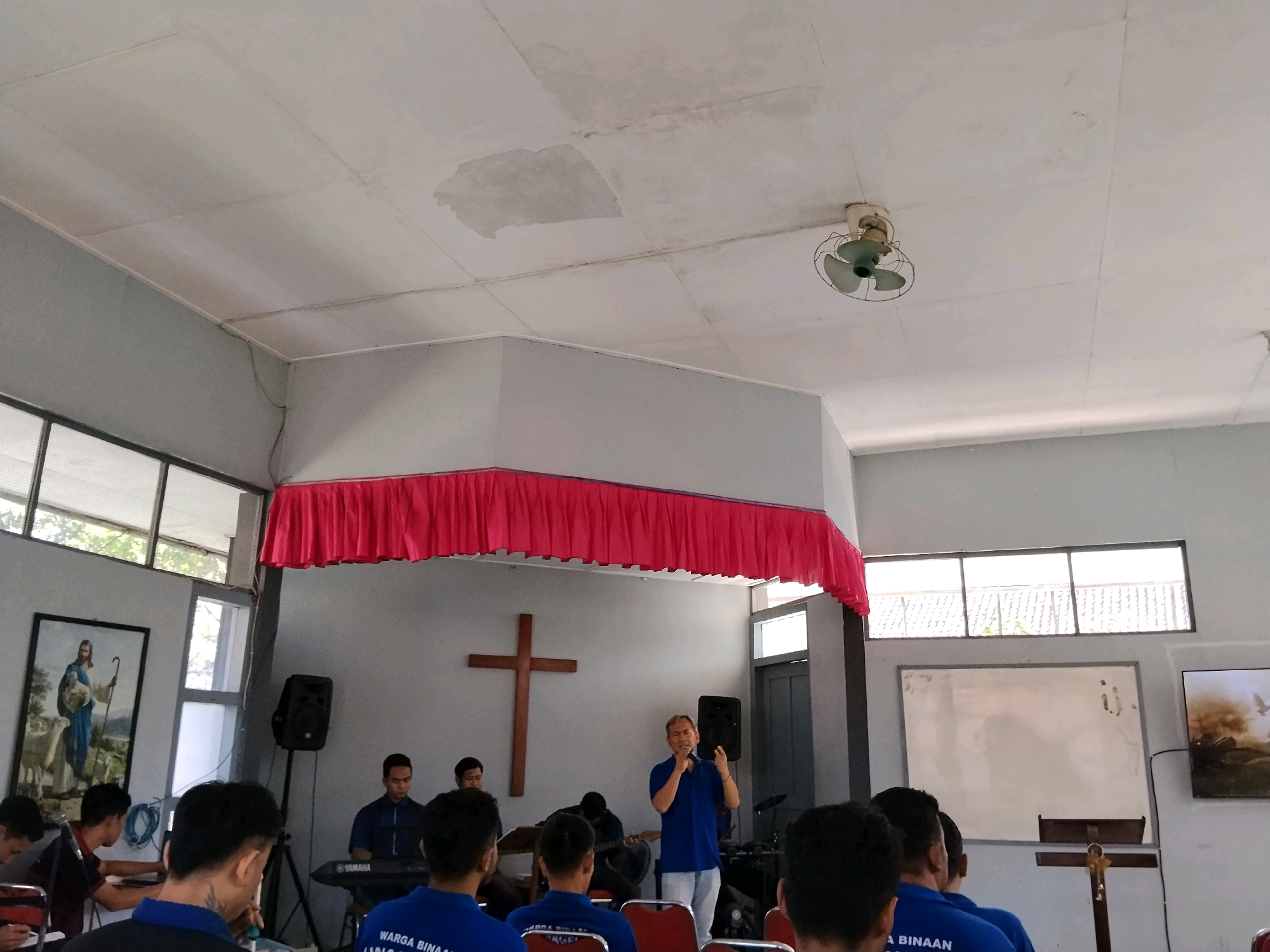 The image depicts an indoor scene, likely a church hall in Magelang, Indonesia. A group of individuals are gathered, with a man in a blue shirt standing and speaking into a microphone, possibly leading a service or presentation. To his left, a wooden cross hangs on the wall. Behind him, a band appears to be set up, with individuals playing keyboards and a guitar. The room features a white ceiling with some water stains and a ceiling fan. A large red draped fabric hangs prominently across the upper portion of the front wall. Windows are visible on the left and right sides of the room. On the left wall, a framed painting of a shepherd with sheep is hung. A whiteboard is present on the right side of the room, and a television screen displays an image on the far right. The attendees are seated in chairs, with some visible in the foreground wearing blue shirts. The overall atmosphere suggests a religious gathering or event.