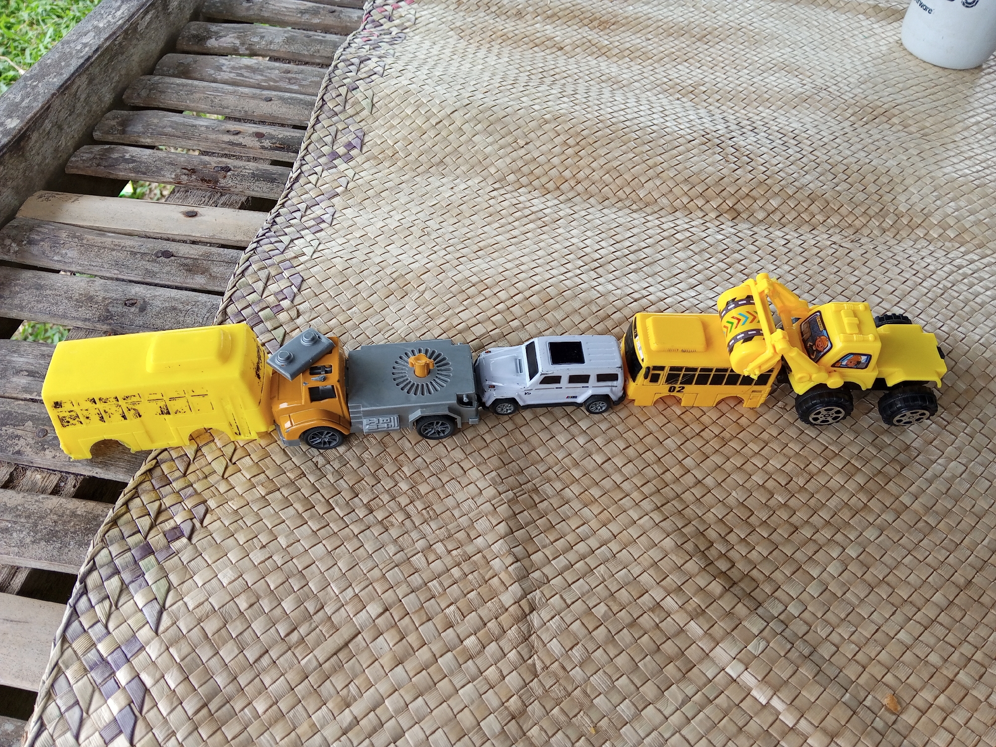 The image shows a line of toy vehicles on a woven mat, which is partially resting on a weathered wooden bench. From left to right, the toy vehicles include a yellow school bus, an orange semi-truck cab, a gray flatbed trailer, a white SUV, another yellow school bus, and a yellow construction excavator. The background suggests an outdoor setting. The image does not contain any people or actions. The location in the city of Palohigeuh, Indonesia is not verifiable from this image alone.